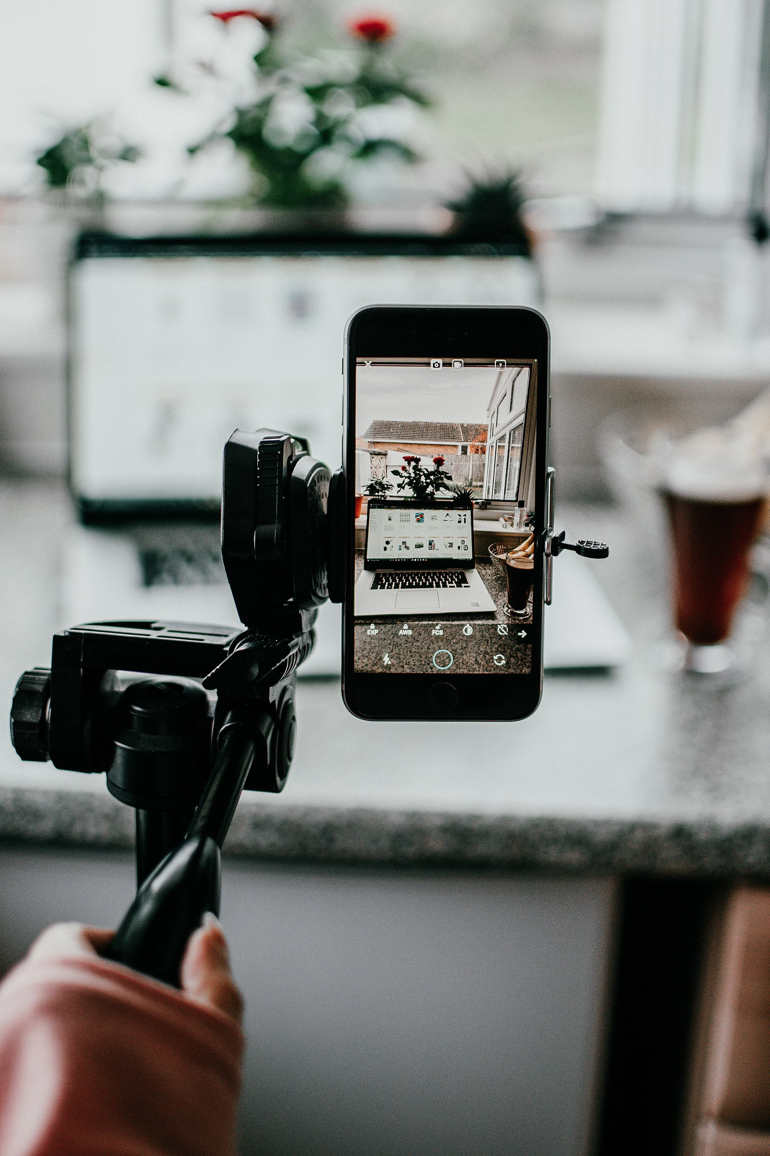 A camera tripod holding a smartphone takes a picture of a laptop on a table, with potted plants and a window in the background.