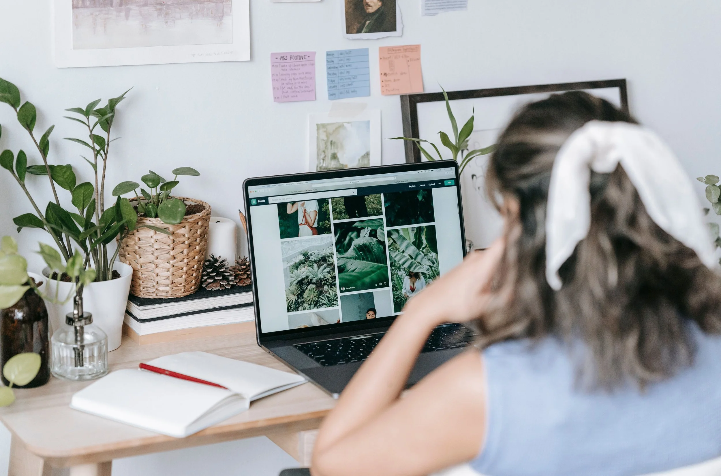 A young girl with curly hair and a large white bow sits at a desk working on a laptop, surrounded by plants, notebooks, and pens, with a white wall decorated with colorful sticky notes and framed pictures in the background.