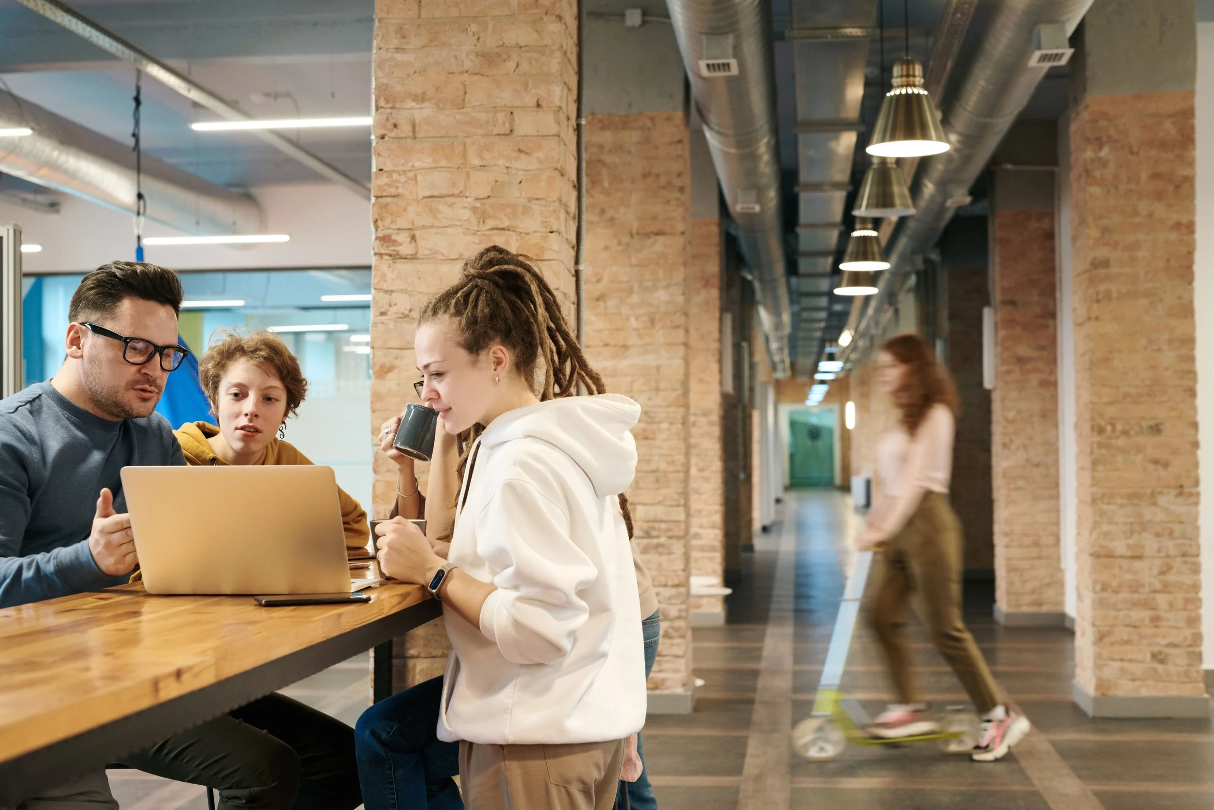 Three people, two women and one young girl, are seated at a wooden table in a modern office or co-working space. The women are engaged in a discussion, with one holding a coffee mug and the other looking at a laptop. The young girl is listening and watching the conversation. The background features exposed brick walls, ductwork on the ceiling, and a woman walking in the corridor.