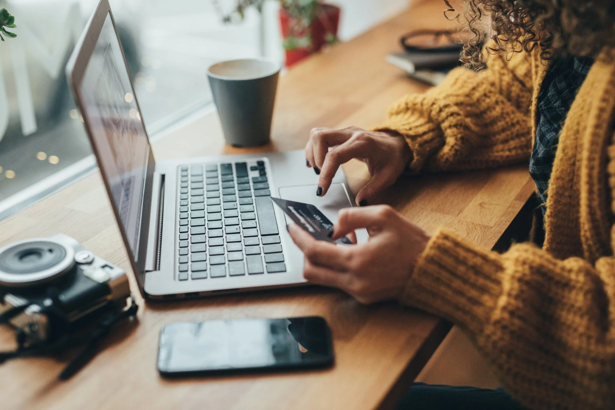 A person with curly hair, wearing a yellow sweater, holding a credit card and typing on a silver laptop at a wooden desk, with a white mug nearby.