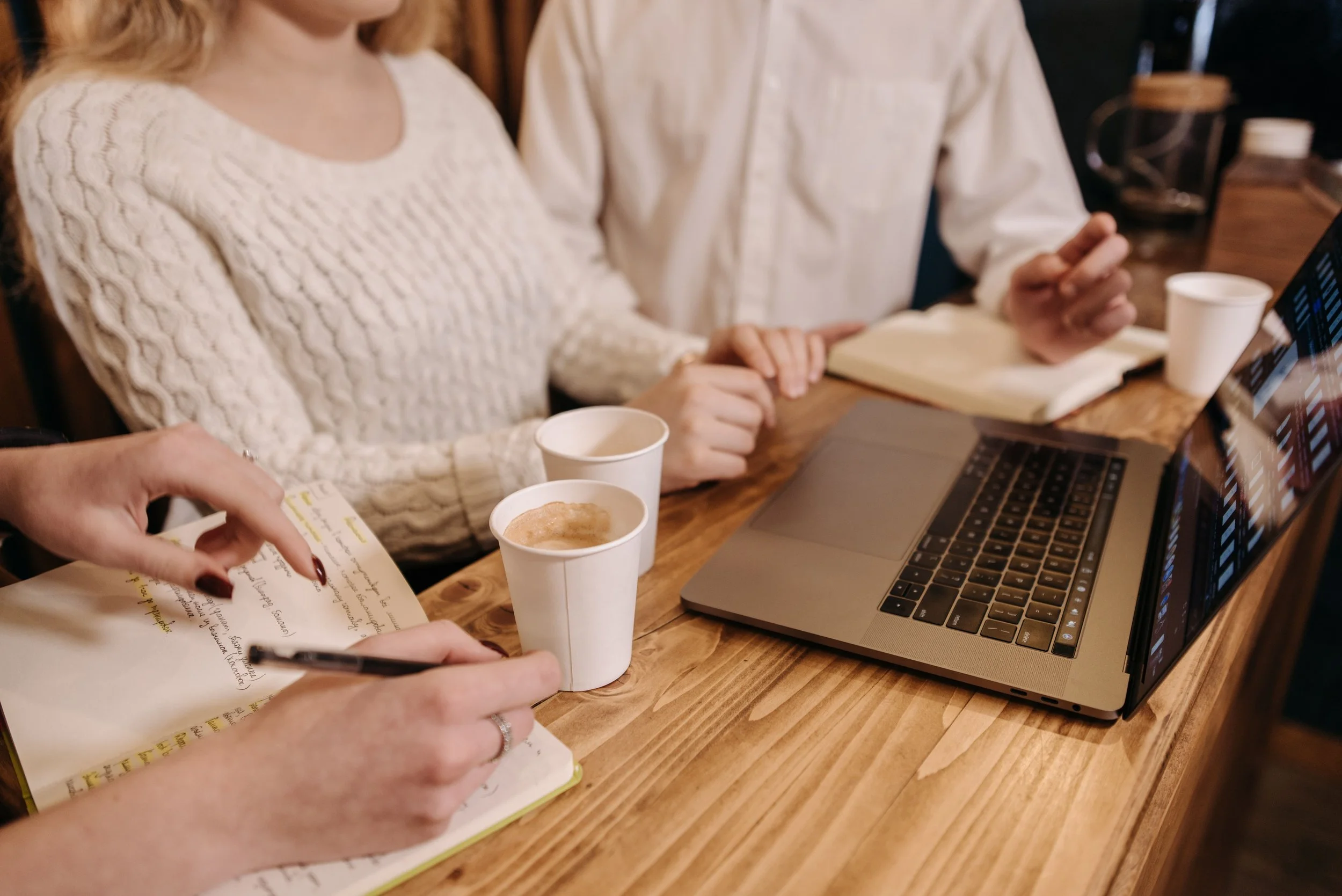 People sitting at a wooden table with a laptop, notebooks, and coffee cups, engaged in discussion or collaboration.