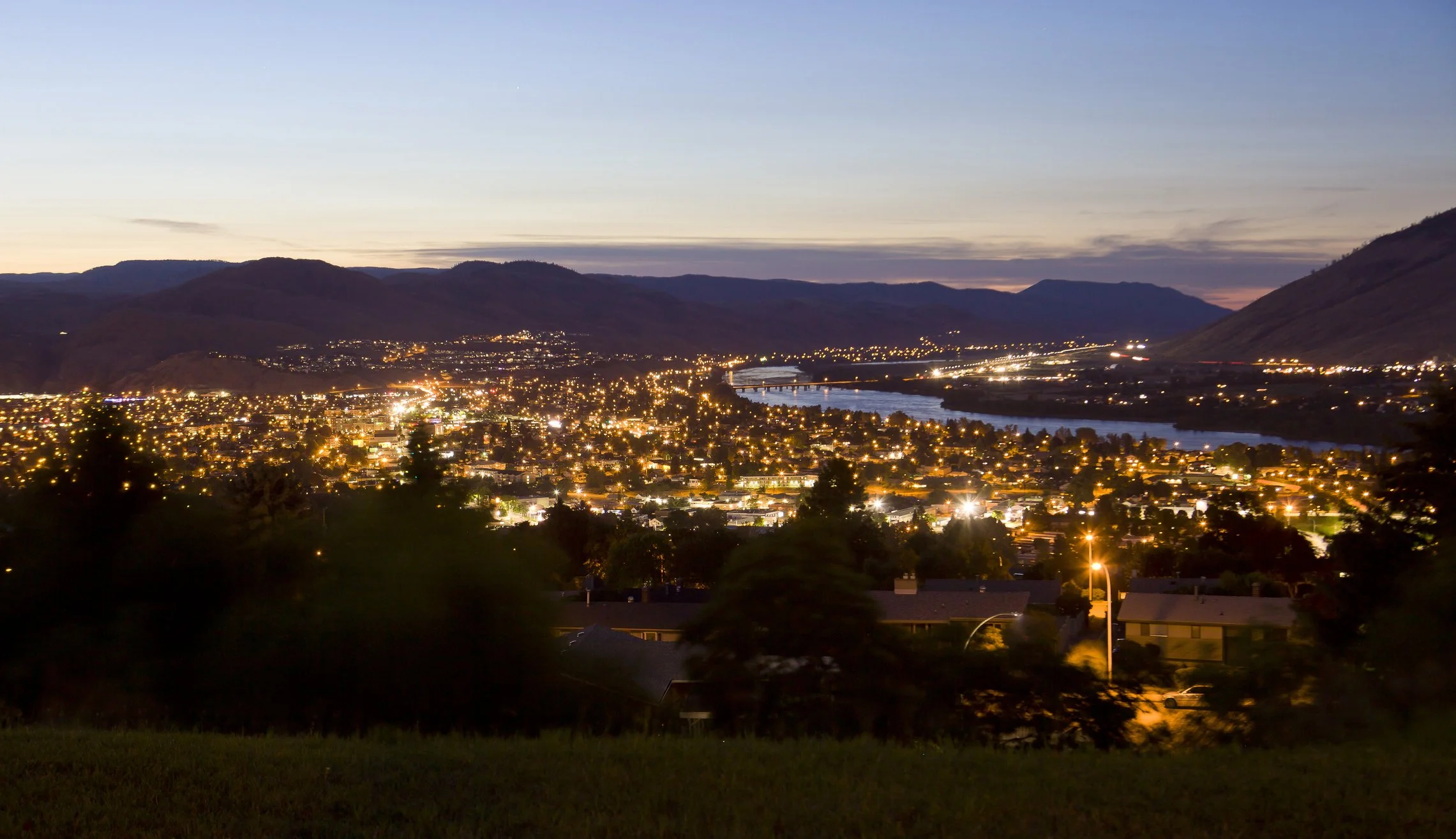 A cityscape at dusk with numerous lights illuminating the town, rolling hills surrounding the city, and a river running through the area.