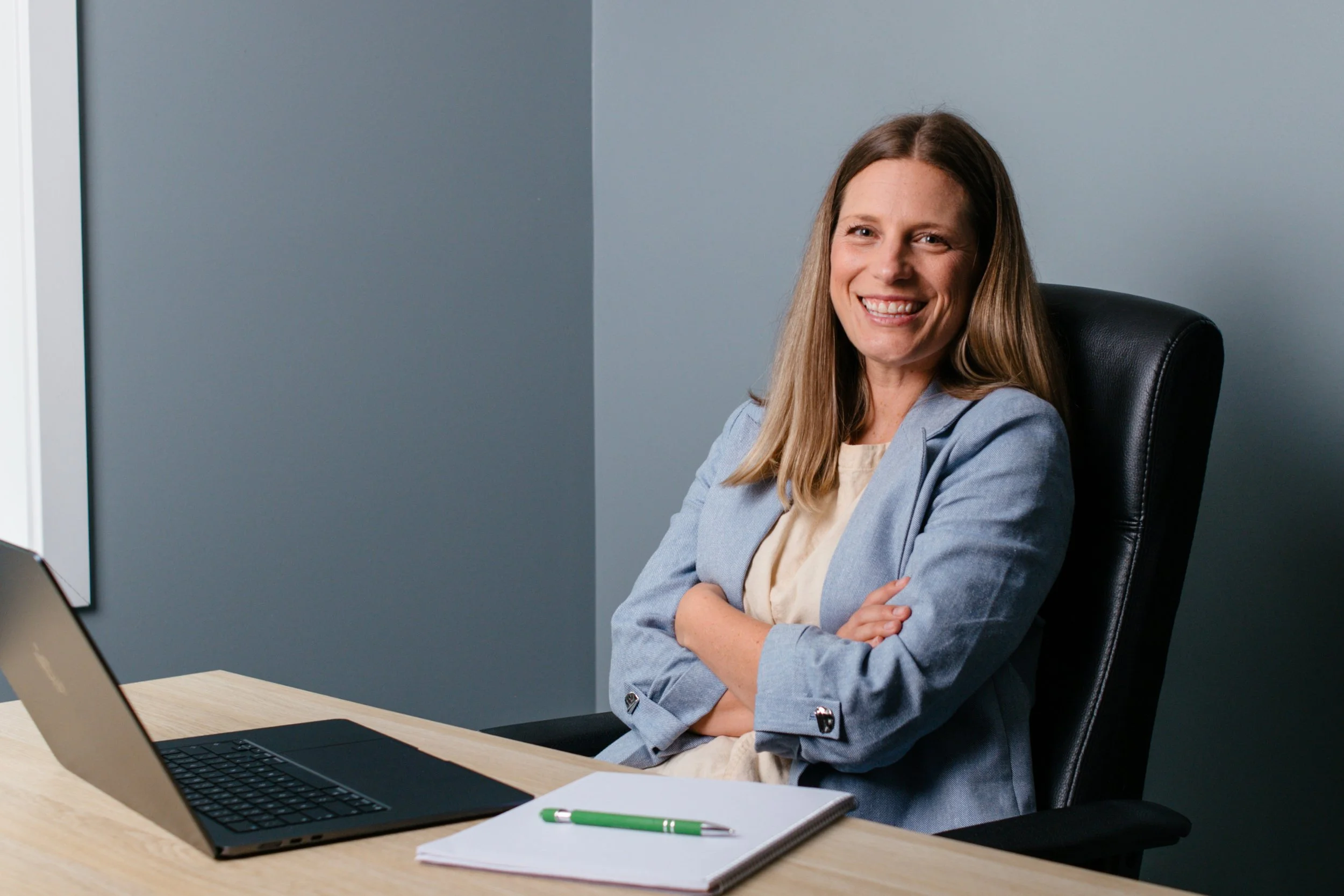 A woman with long brown hair smiling, sitting in a black office chair with arms crossed, in front of a desk with a laptop, a notebook, and a pen, against a gray wall.