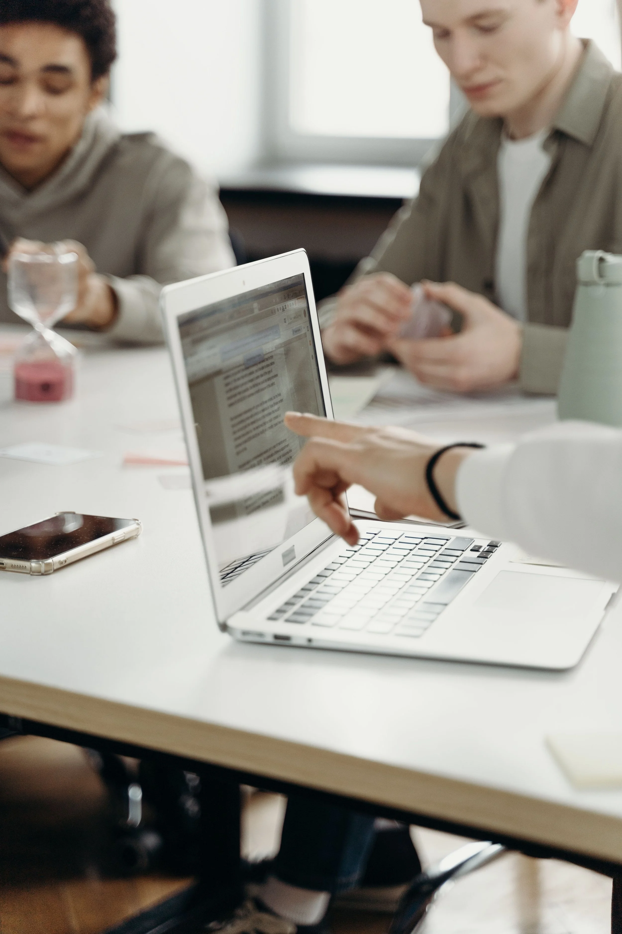 Two people are sitting at a table, working on laptops with papers and a phone nearby. One individual is pointing at the laptop screen. The scene suggests a collaborative work or study session in a bright room.