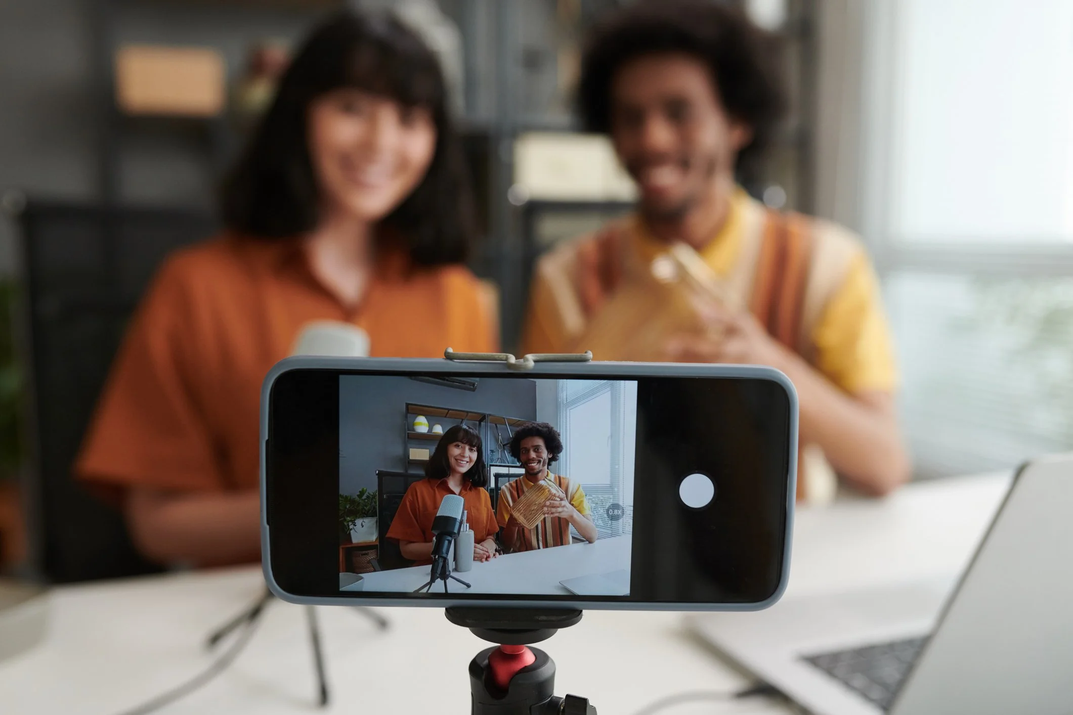 A smartphone on a tripod captures a photo of a woman and man sitting at a table, smiling, with a bookshelf and window in the background