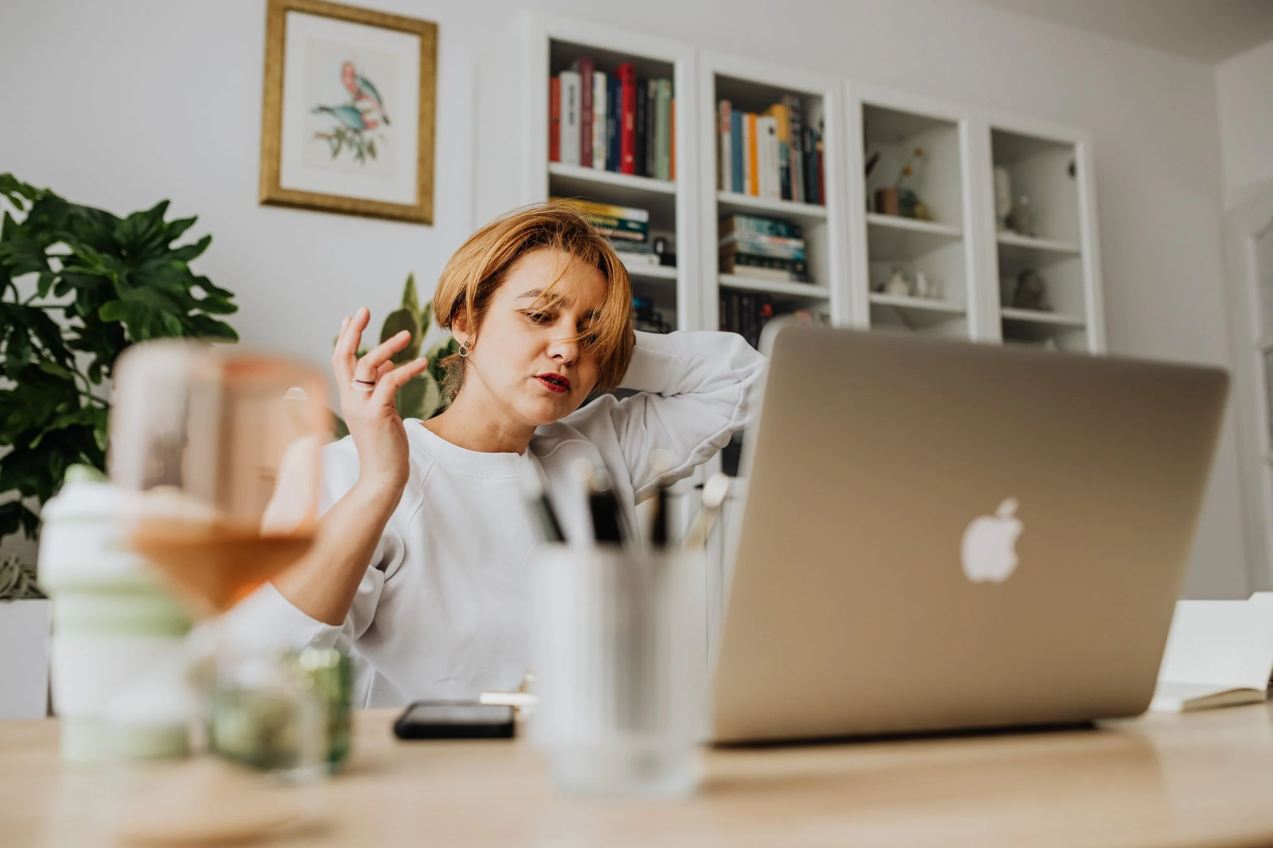 A woman with short red hair and earrings sitting at a desk, working on a silver laptop in a home office with white bookshelves filled with books behind her. There are houseplants and various items on the desk.