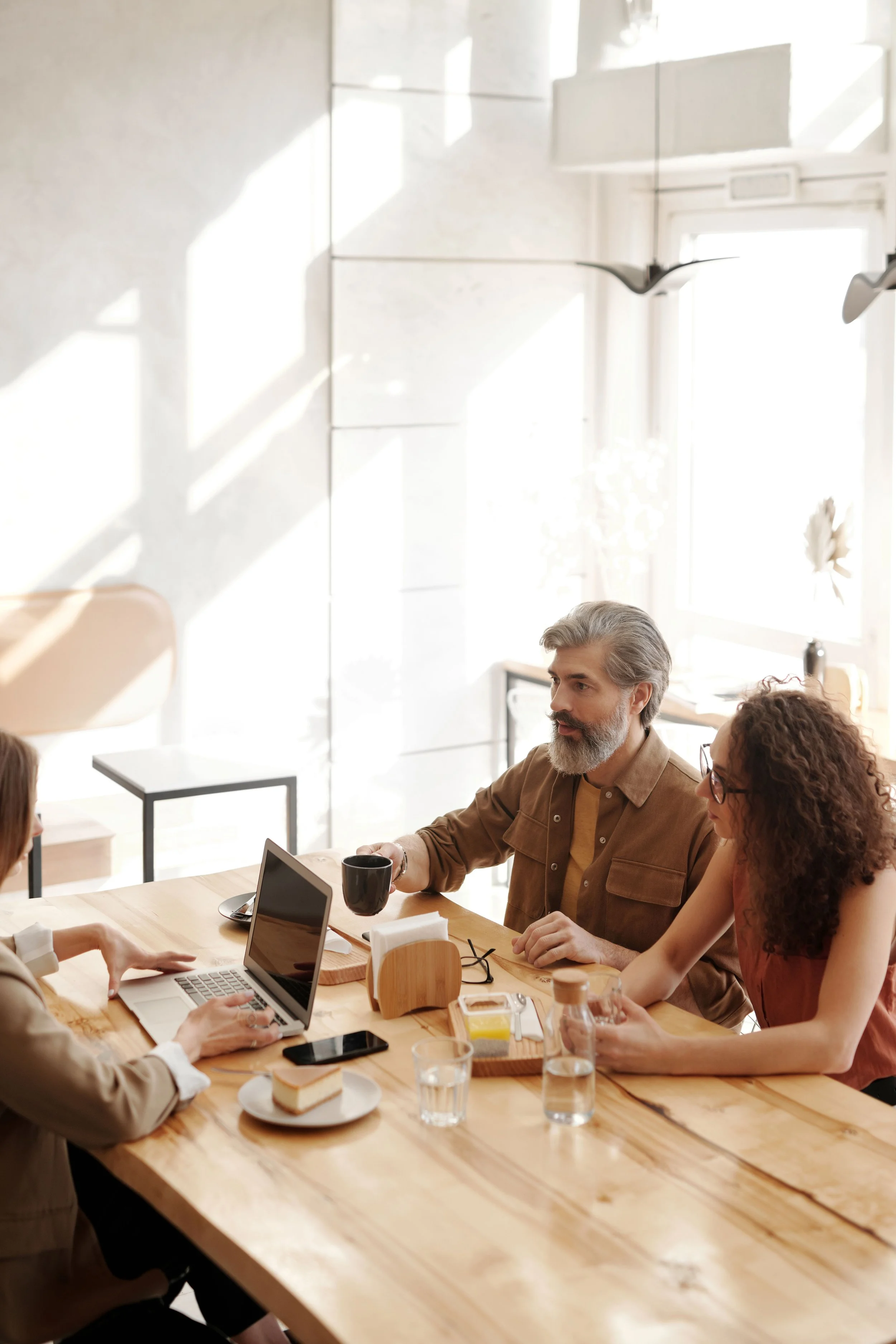 A group of people gathered around a wooden table in a bright room, engaging in a discussion or meeting. There are laptops, coffee cups, glasses of water, and a slice of cake on the table. Sunlight streams through the windows.