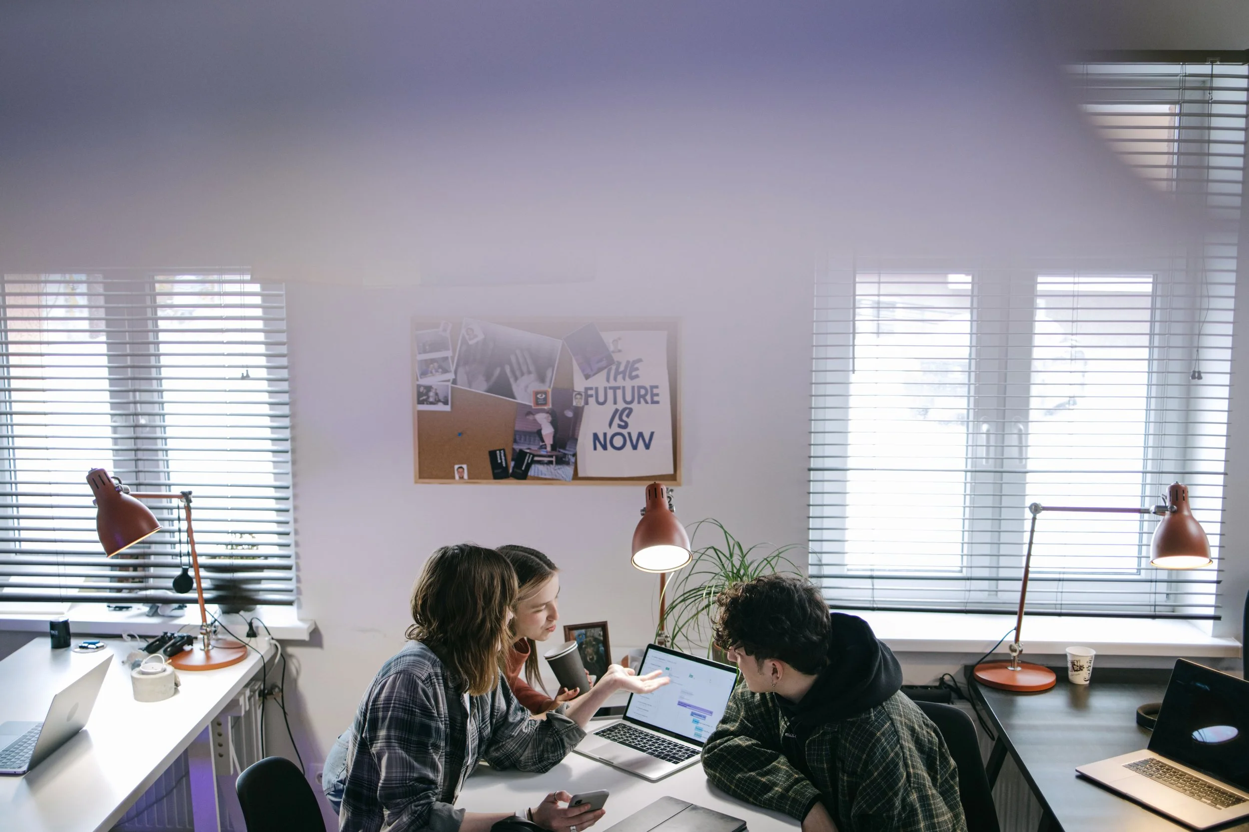 People sitting at a table in a bright office, engaged in conversation with laptops, notebooks, and a smartphone on the table. Large windows with blinds are in the background, and a bulletin board with a motivational poster.