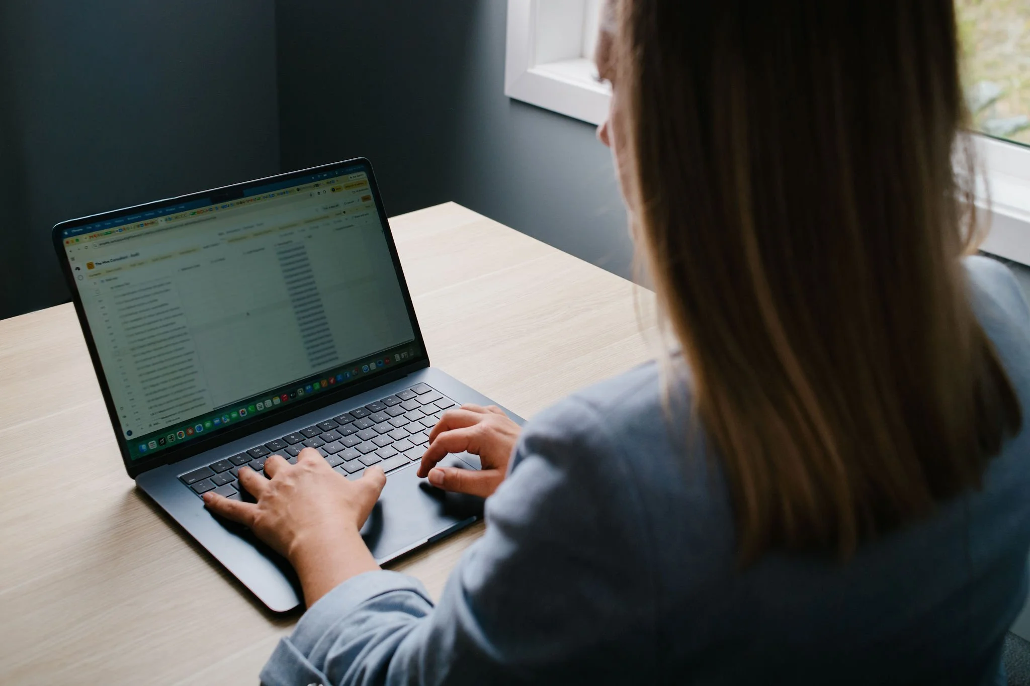 A person with brown hair is sitting at a light wood desk, typing on a silver laptop with a black keyboard, near a window with white trim.