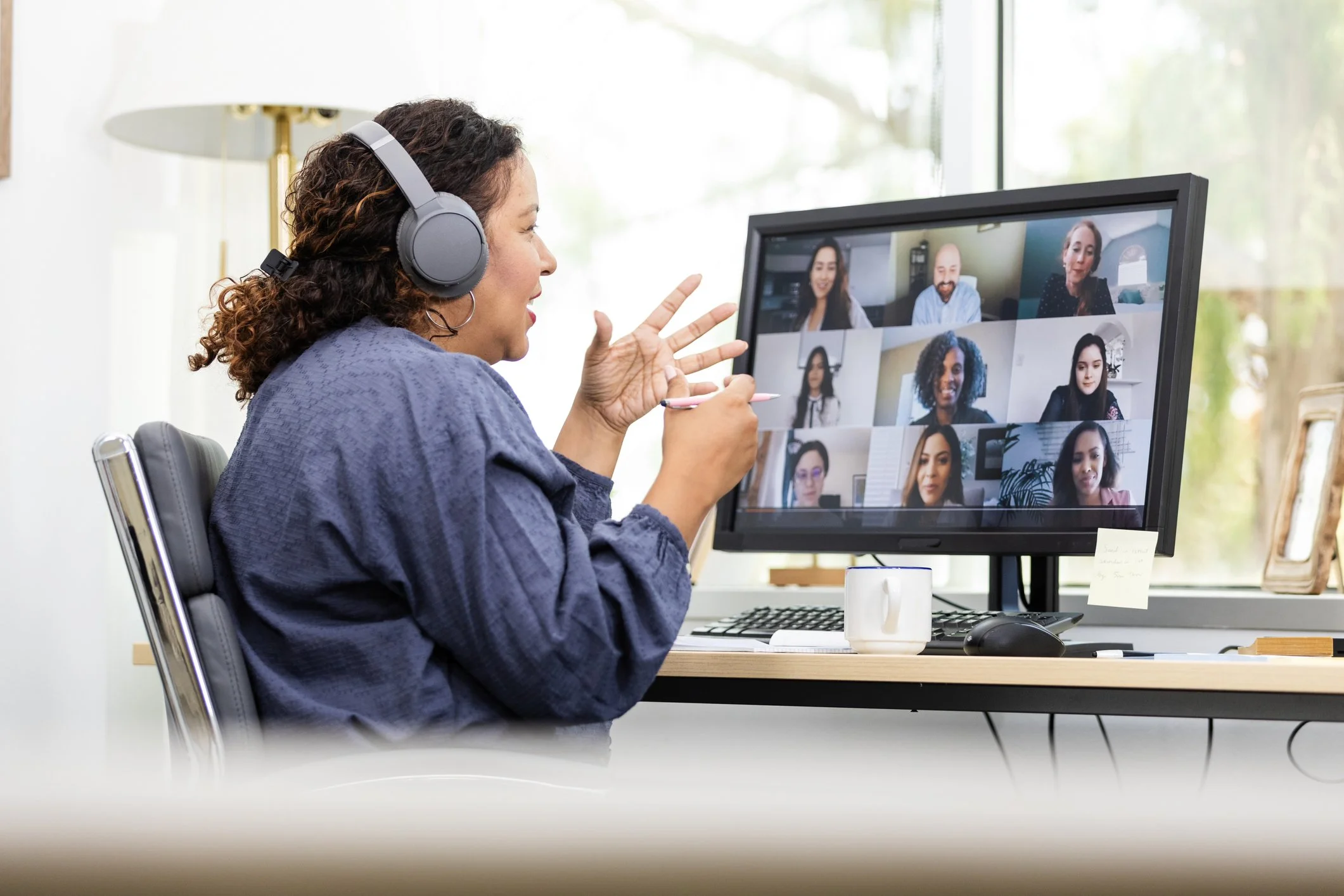 Woman with curly hair wearing headphones participating in a virtual meeting on her computer, with multiple people visible on the screen.