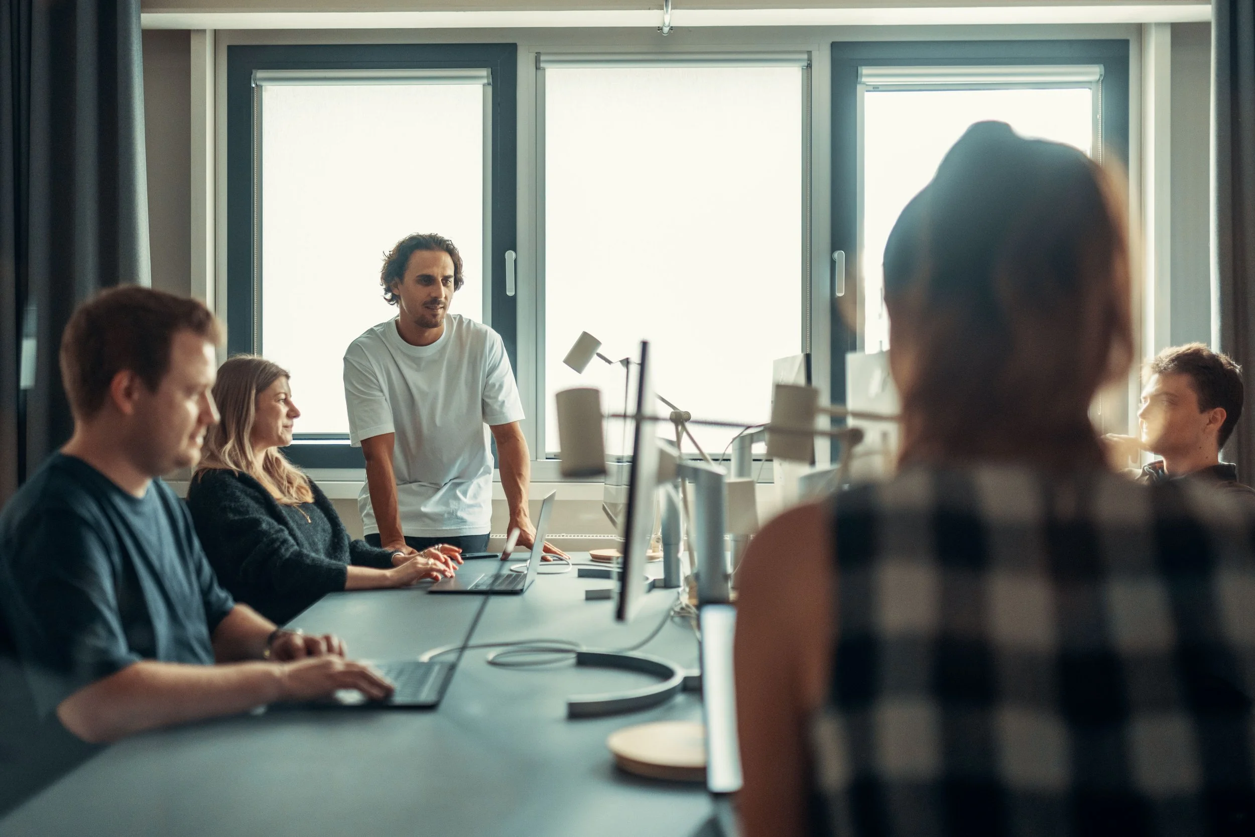 A group of people sitting at a long conference table with laptops, while a man stands talking. They are in a modern office with large windows and natural light.