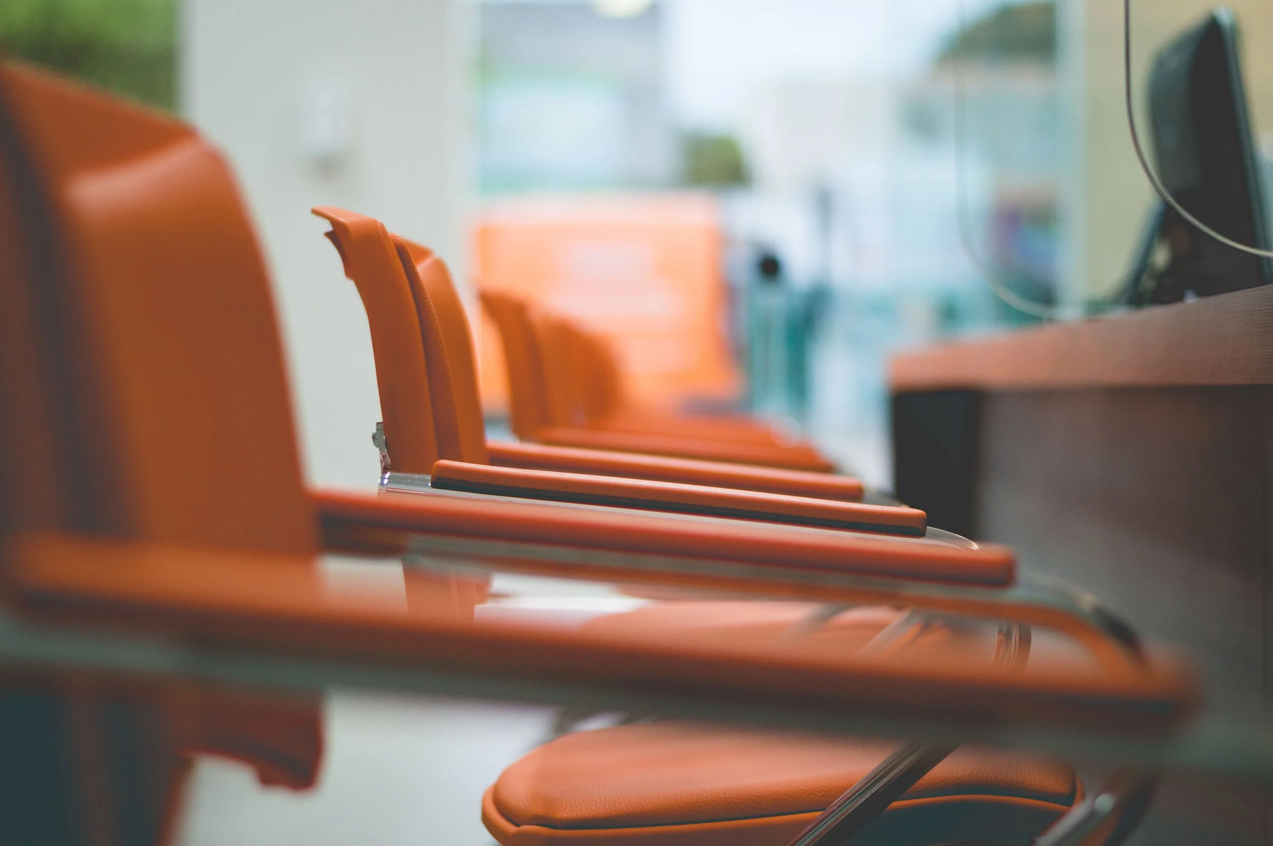 A row of empty orange chairs with chrome legs next to a desk with a computer monitor, in a modern office or waiting area.