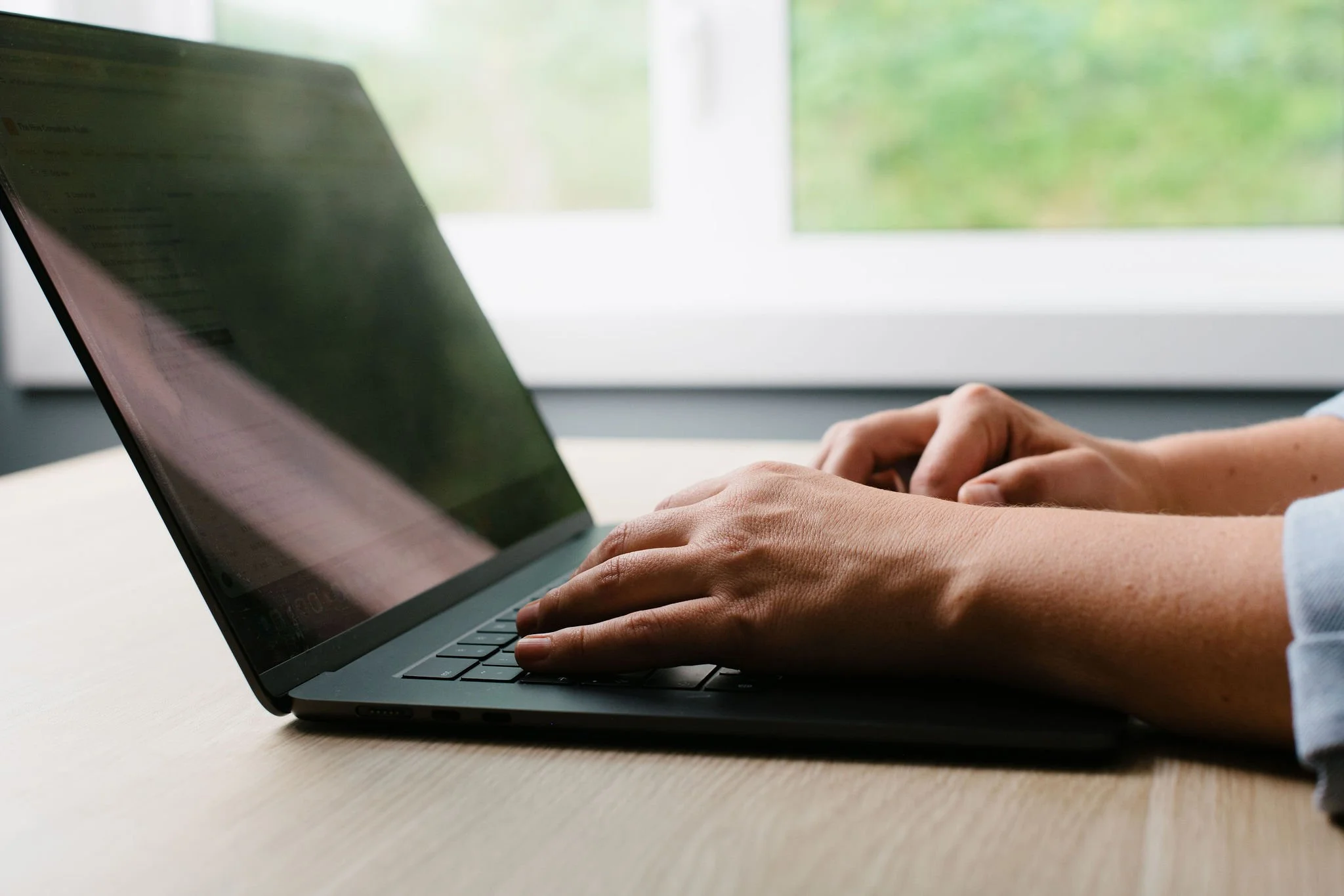 Close-up of hands typing on a laptop keyboard, with a window showing green foliage in the background.