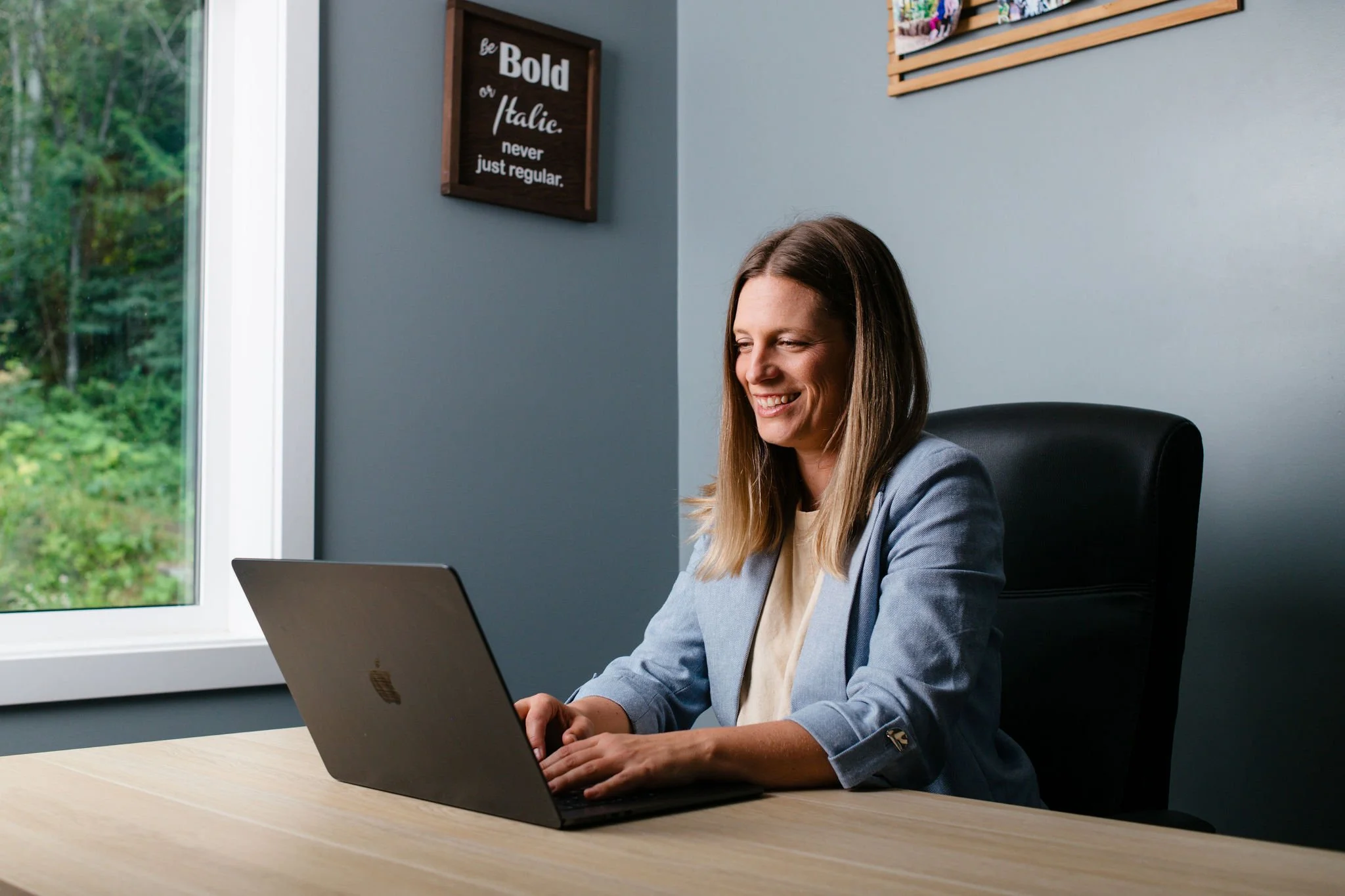 A woman with brown hair smiling as she works on a laptop at a desk in an office, with a window showing green trees outside. There is a motivational sign on the wall that says, "Be bold or italic. never just regular."