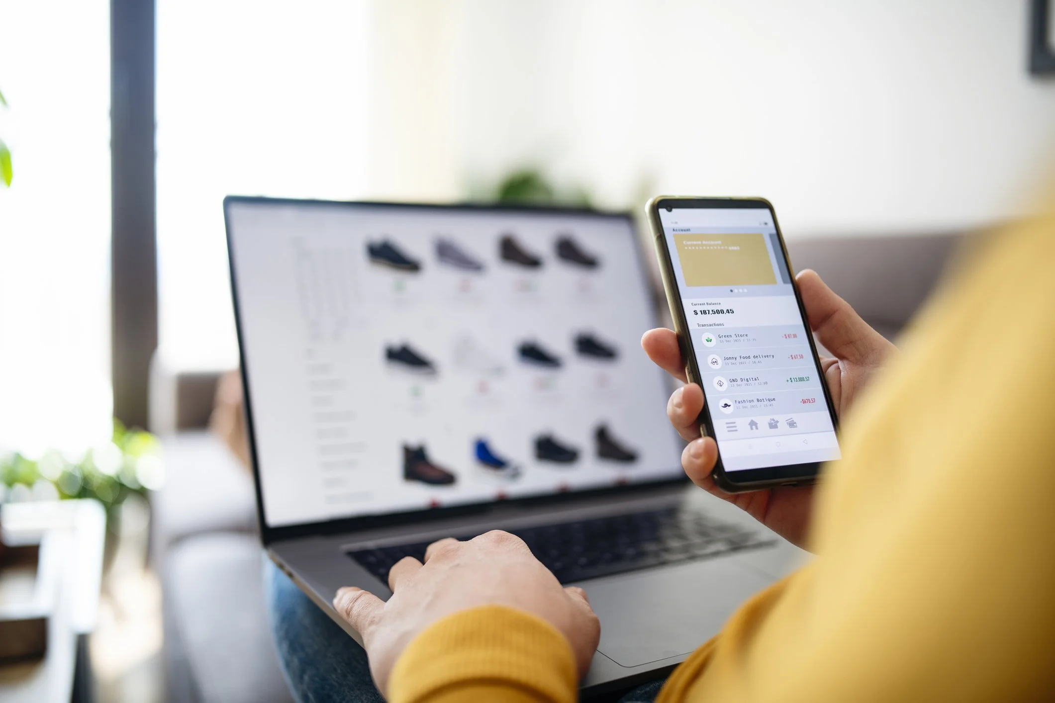 A person wearing a yellow sweater holding a smartphone with a financial app open, showing account balance and transactions, while using a laptop with a website displaying sneakers in the background.