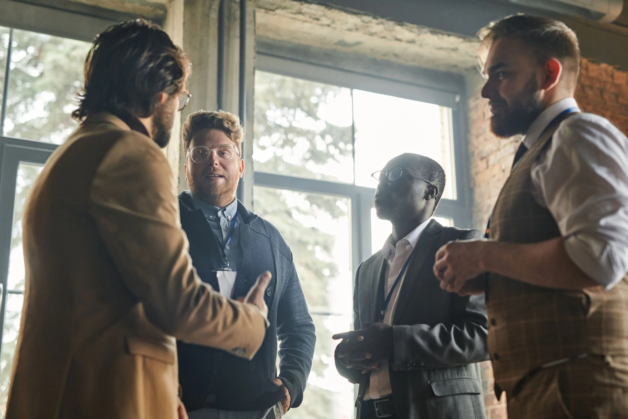 A group of five men engaged in a conversation at an indoor setting with large windows and exposed brick walls, some wearing glasses and business attire.