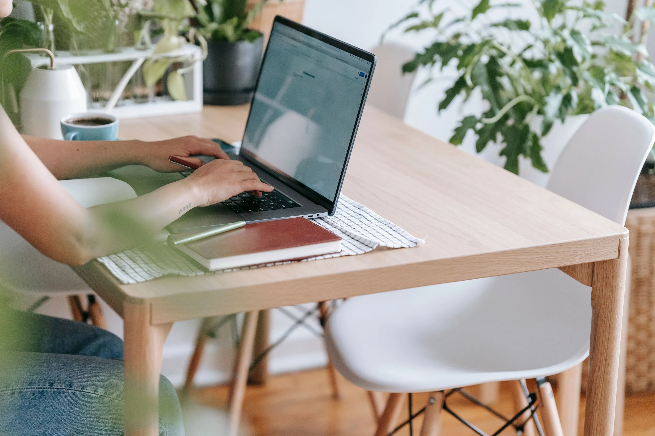 Person working on a laptop at a wooden dining table with a coffee mug, a notebook, and houseplants in the background.
