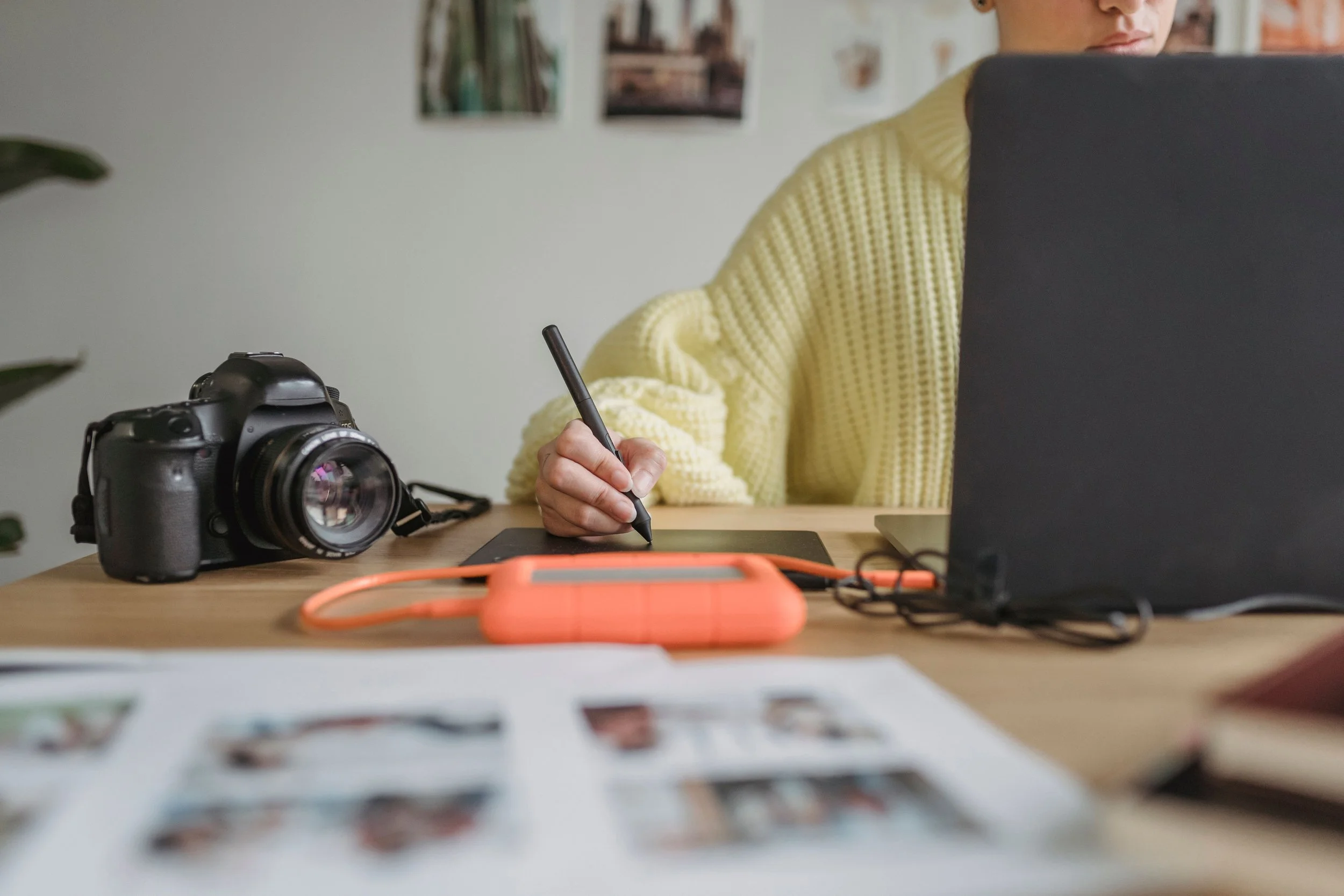 Person sitting at a desk using a graphics tablet and stylus, with a camera, external hard drive, and photo contact sheet on the desk.