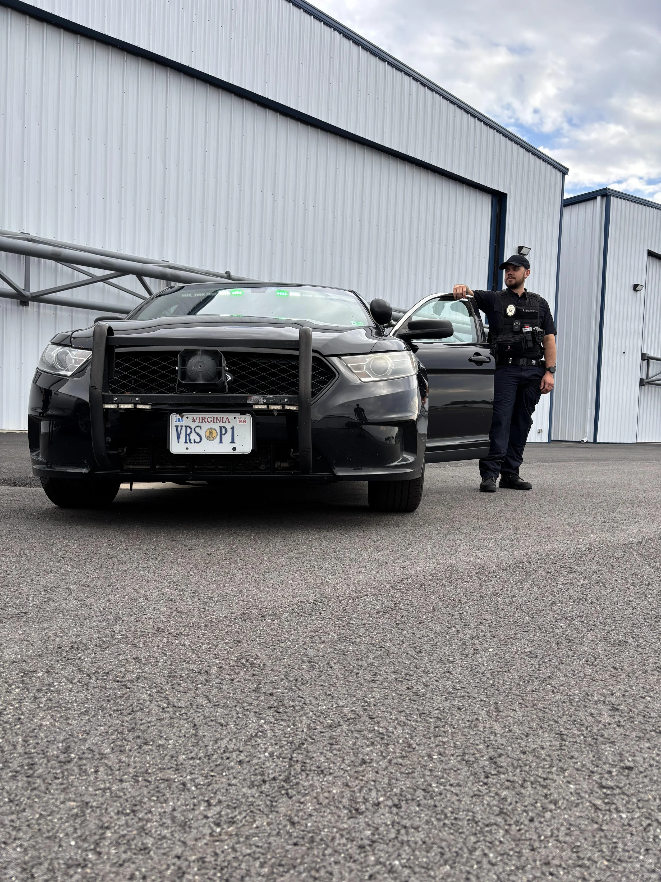 A police officer standing next to a black police car parked in front of a large industrial building. The car has a push bumper on the front and green lights on the dashboard. The officer is wearing a black uniform with patches and a hat. The sky is cloudy.