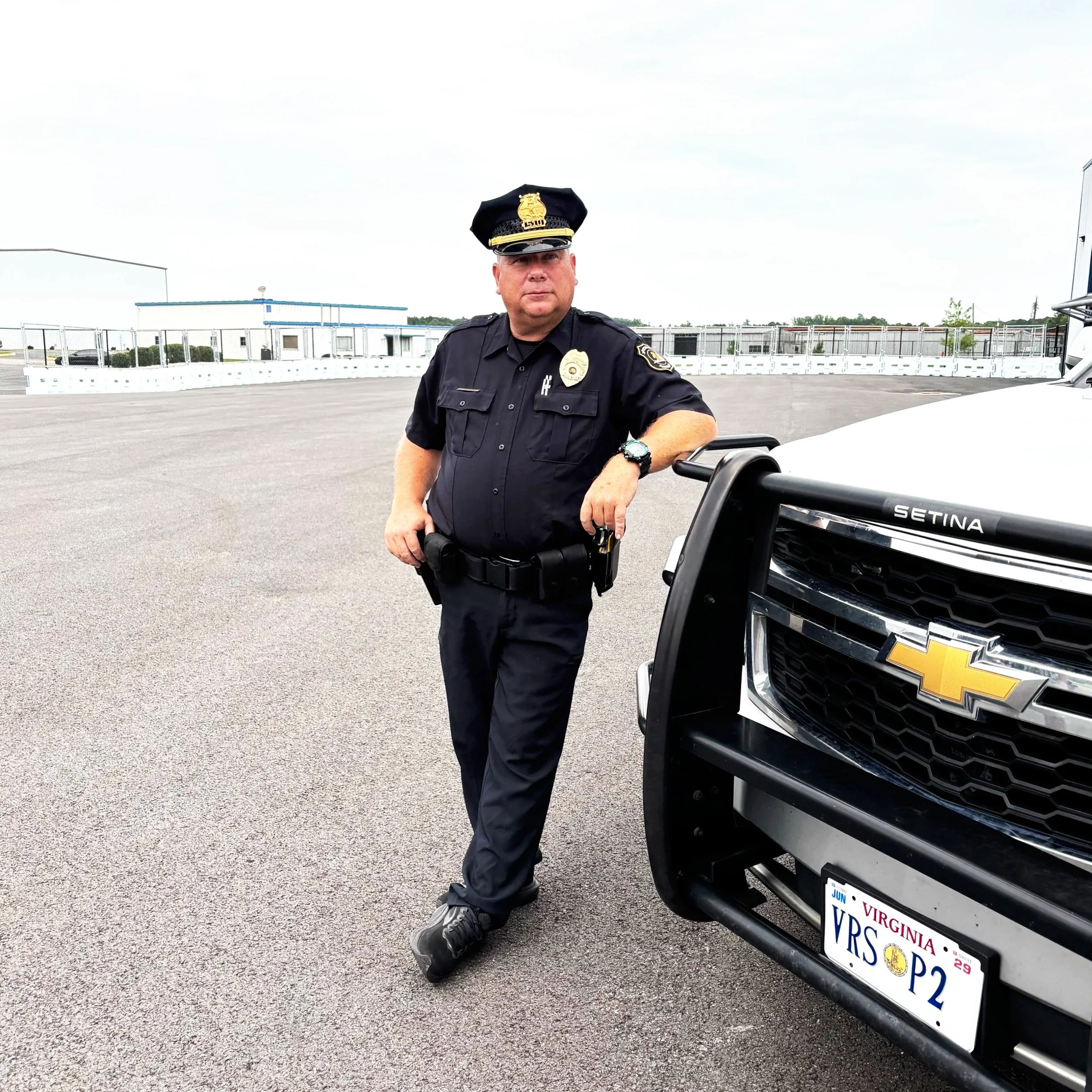A police officer stands next to a black Chevrolet vehicle in a parking lot, with buildings and a chain-link fence in the background, under a cloudy sky.
