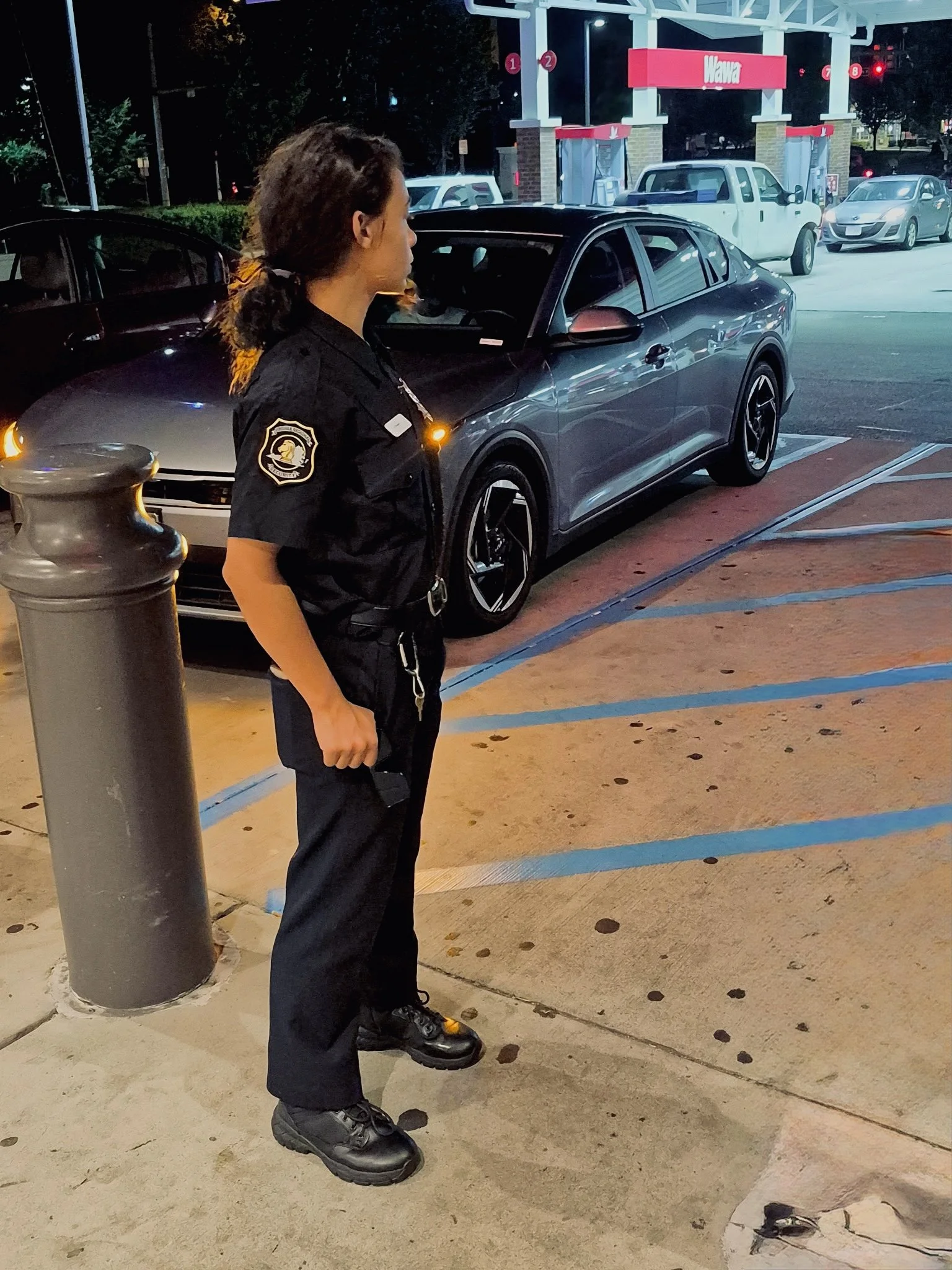 A security guard standing outside at night near a parking lot with multiple cars, including a gray sedan and a pickup truck. The guard is wearing a black uniform with a badge and is looking to the right. The background features a Wawa gas station and illuminated red lights.