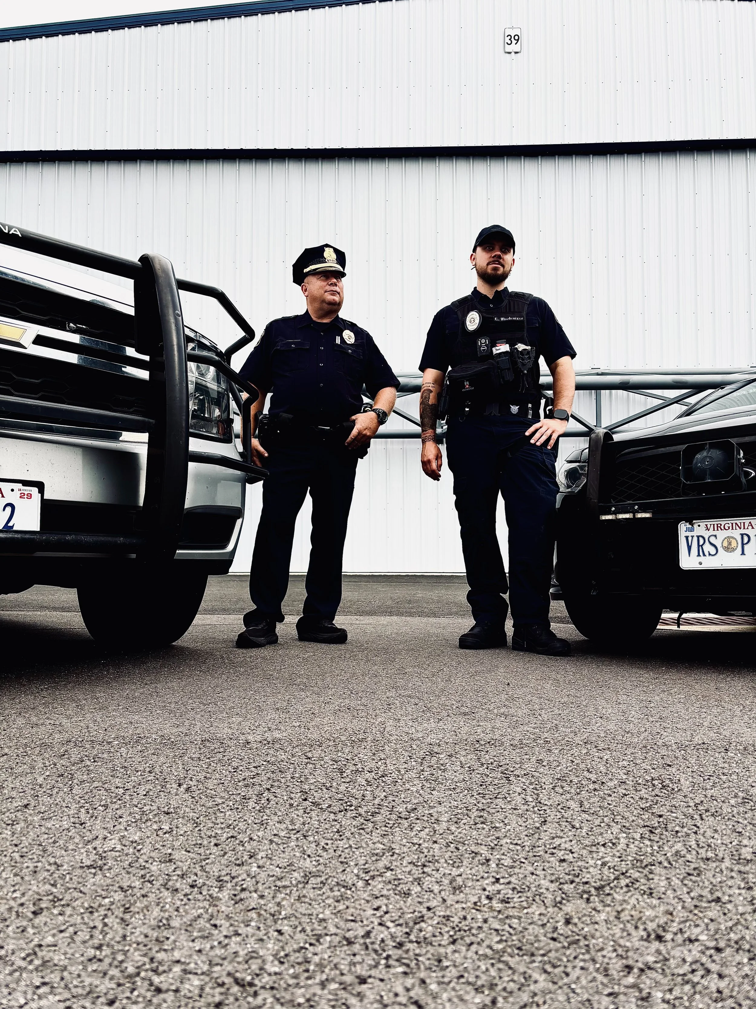 Two police officers standing in front of vehicles, one male police officer in a black uniform and hat with a badge, and a younger male officer with tattoos wearing a vest and black cap, with a white industrial building in the background.