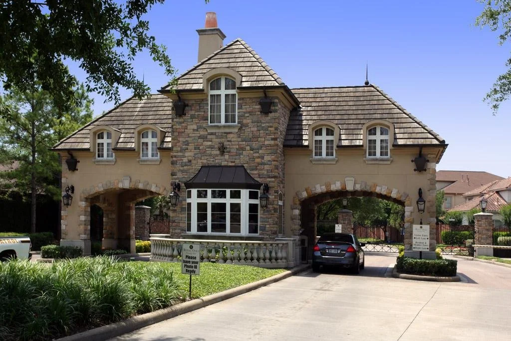 Residential gated community entrance with a stone and stucco gatehouse, archways for vehicles, a black gate and a parked car, surrounded by landscaped greenery and trees.