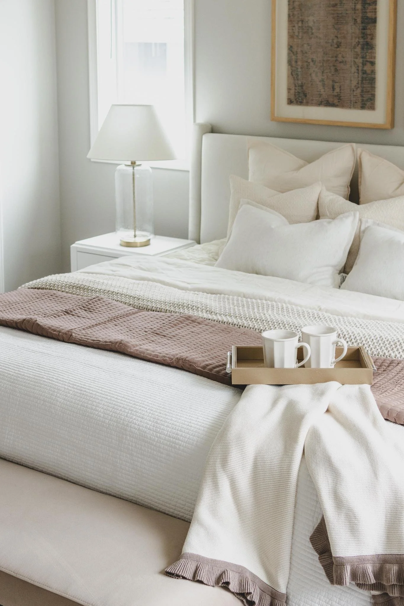 A neatly made bed with cream-colored bedding, beige and white pillows, a pinkish-brown textured blanket, and a tray with two white mugs and a cream cloth, beside a nightstand with a glass lamp, in a bright room with a window and framed artwork.