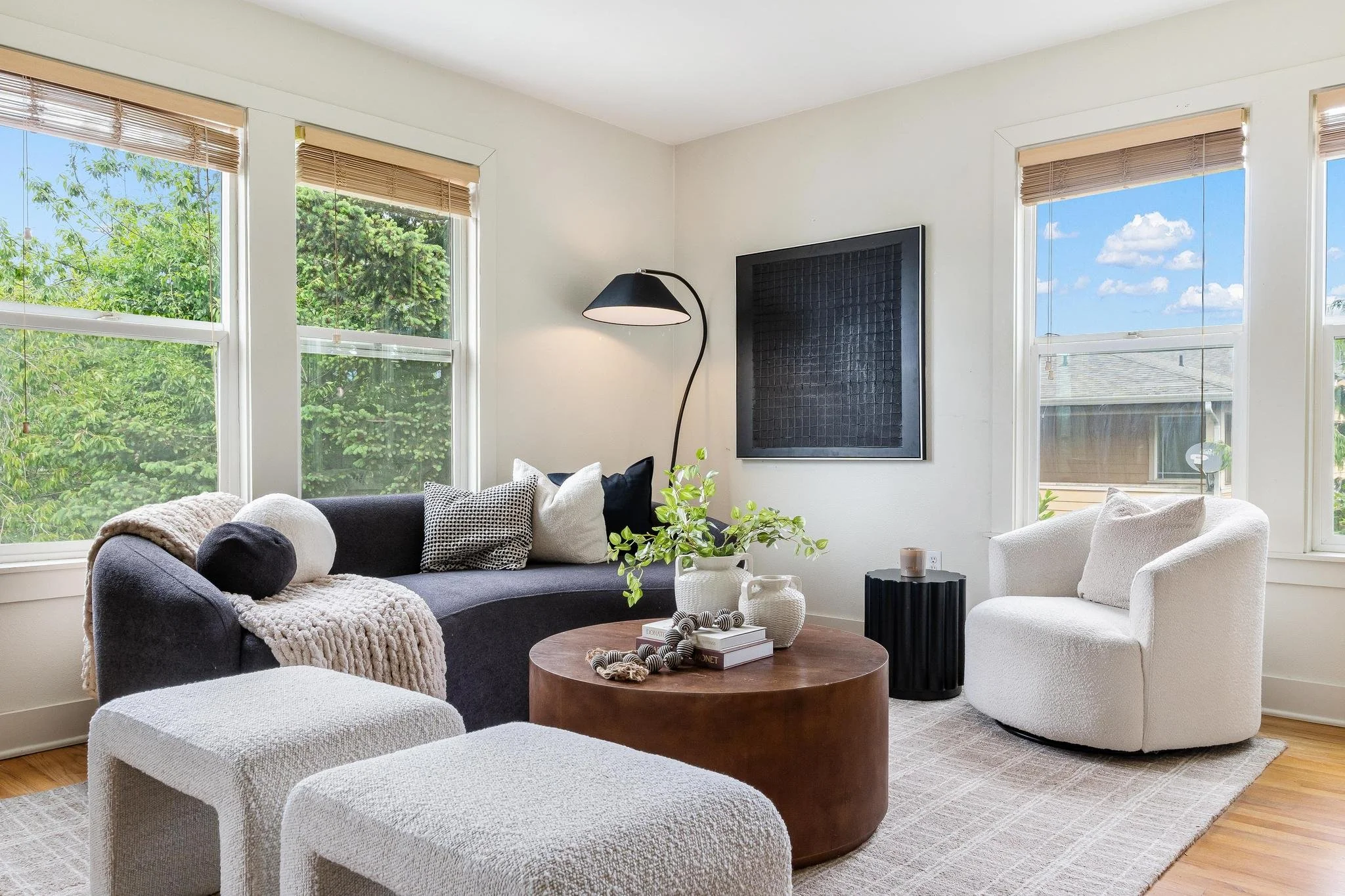 Bright living room with large windows, white walls, a black wall clock, gray couch with pillows, white armchair, round wooden coffee table with decorative items, and lush green trees outside.