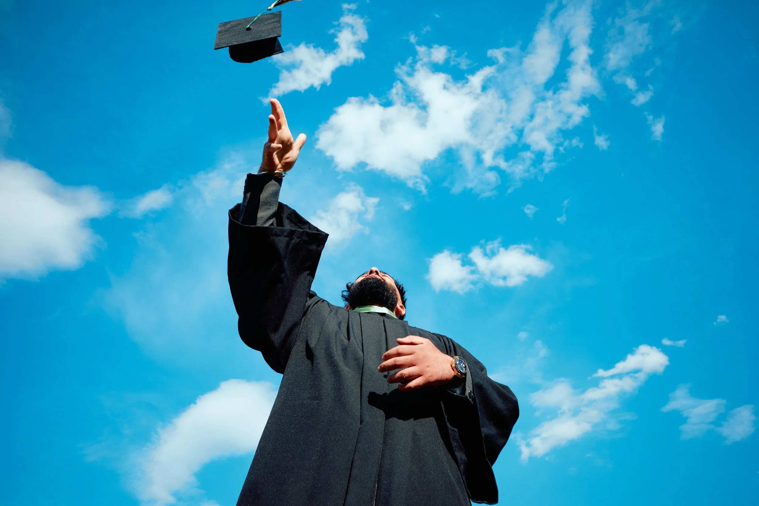 A graduate in a black cap and gown tossing their cap into the blue sky with white clouds, viewed from below.