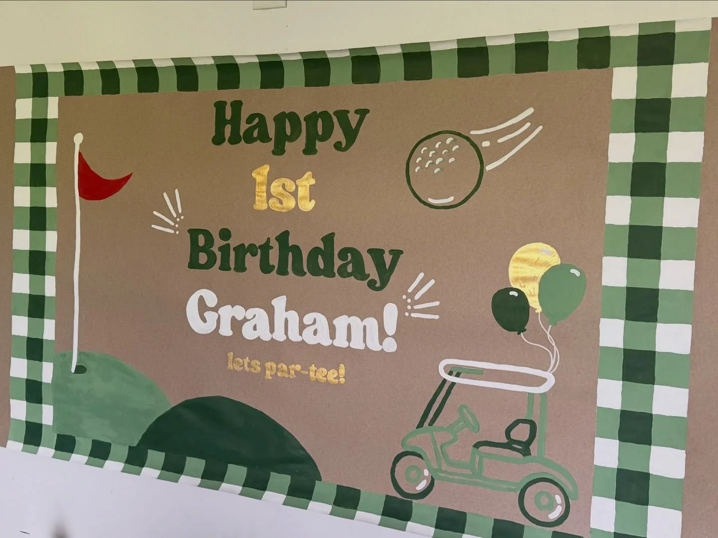 banner with a gingham border that says happy 1st birthday graham! with a golf cart and balloons wiht a flying golf ball and a red flag with the green of where to putt the ball.