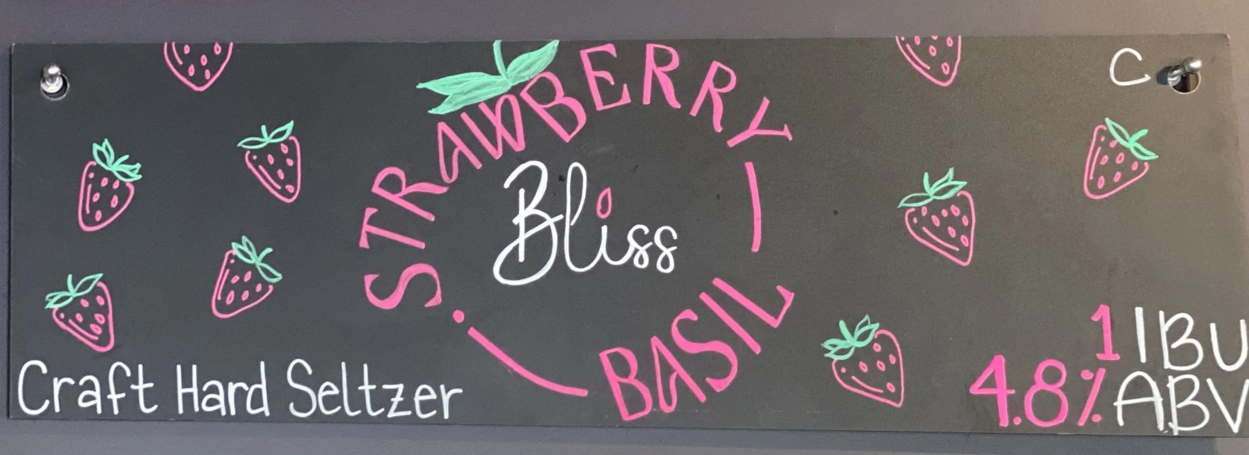 Chalk design that says Strawberry Basil. The words are in the shape of a strawberry. With a leaf in green above, in the middle says Bliss. All around are little strawberries in pink and green. Below says craft hard seltzer. 