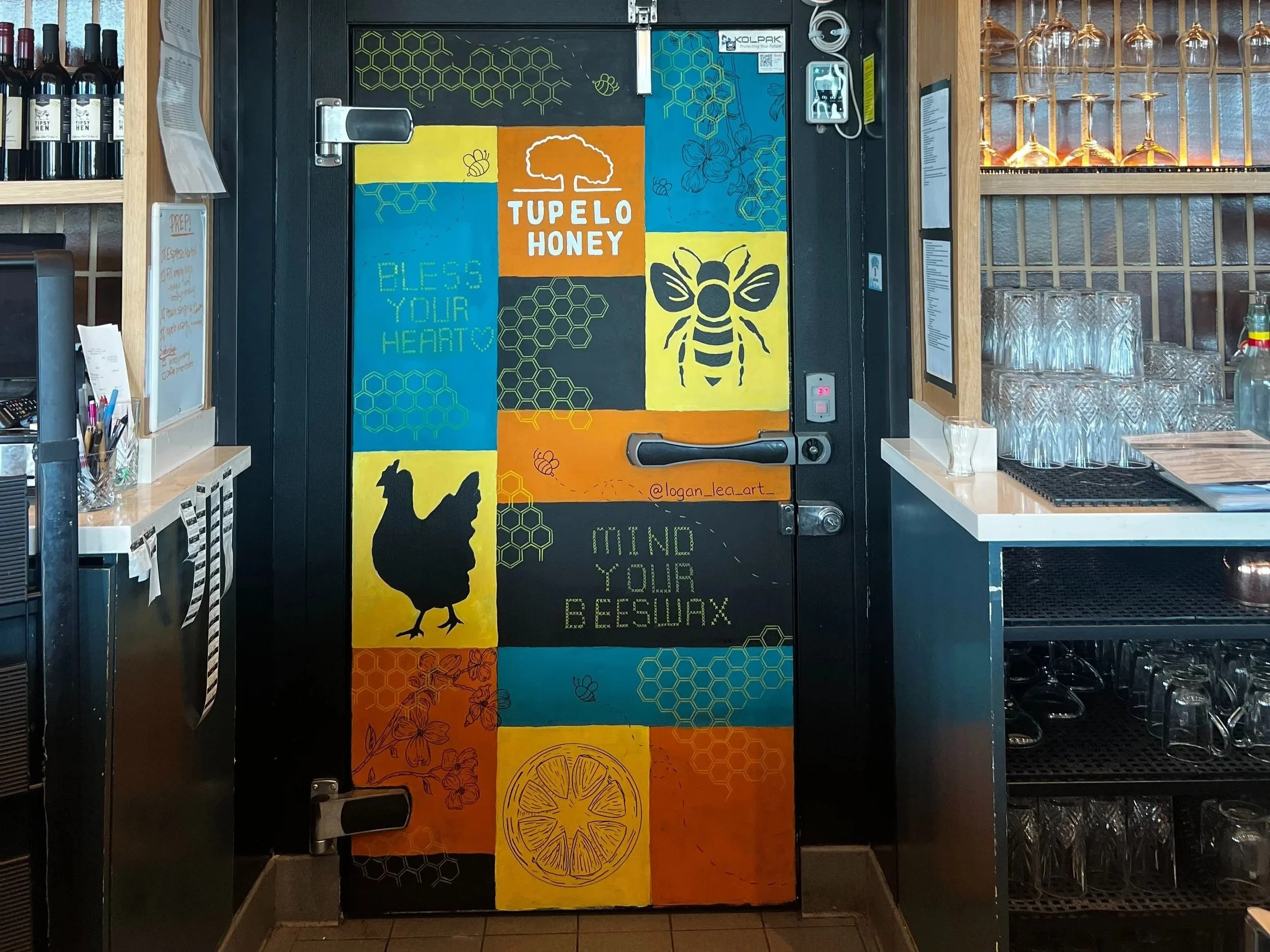 image of a mural at a bar on the cooler walk in door. has yellow orange blue and black squares with a drawing of a chicken, bee, flowers, and lemon. Says tupelo honey and has honeycomb drawings on it. says mind your beeswax and bless your heart on it