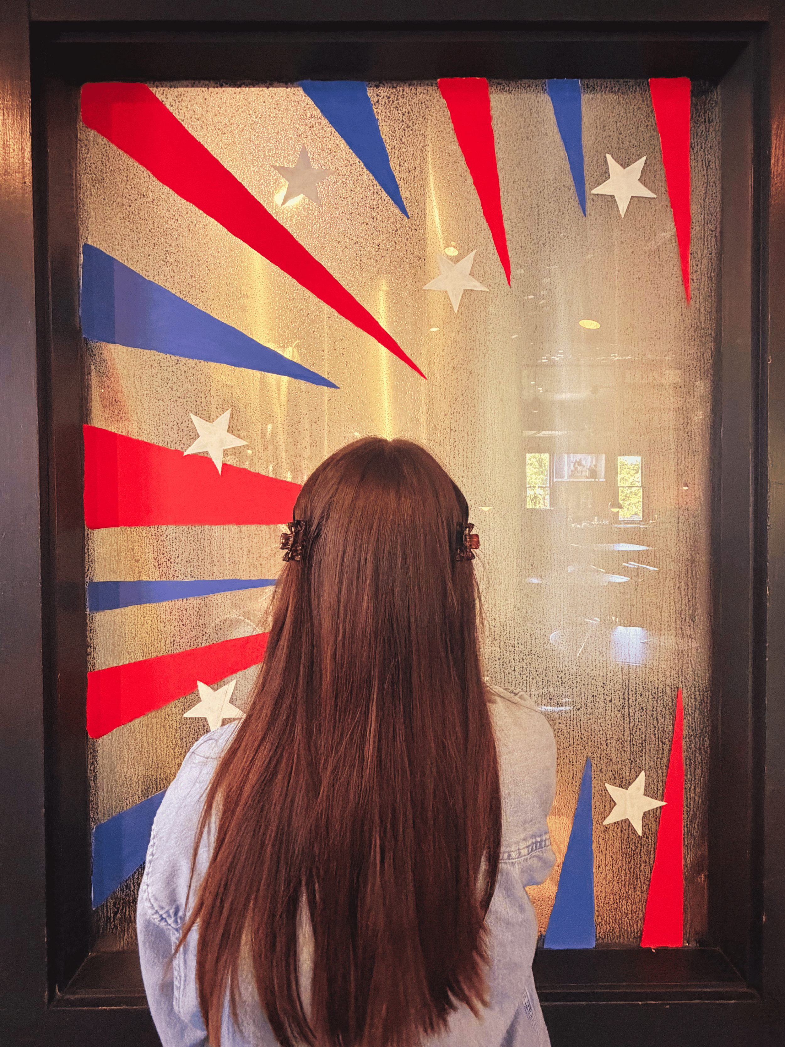 woman with long brown hair standing infront of a window with a painting of stars and stipes in red white and blue with the title at the top says colossal art by logan in green