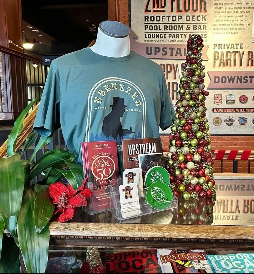 t-shirt on a mannequin in forest green that says ebenezer with a drawing of silhouette of a man in a top hat holding a candle and says winter warmer beside him.