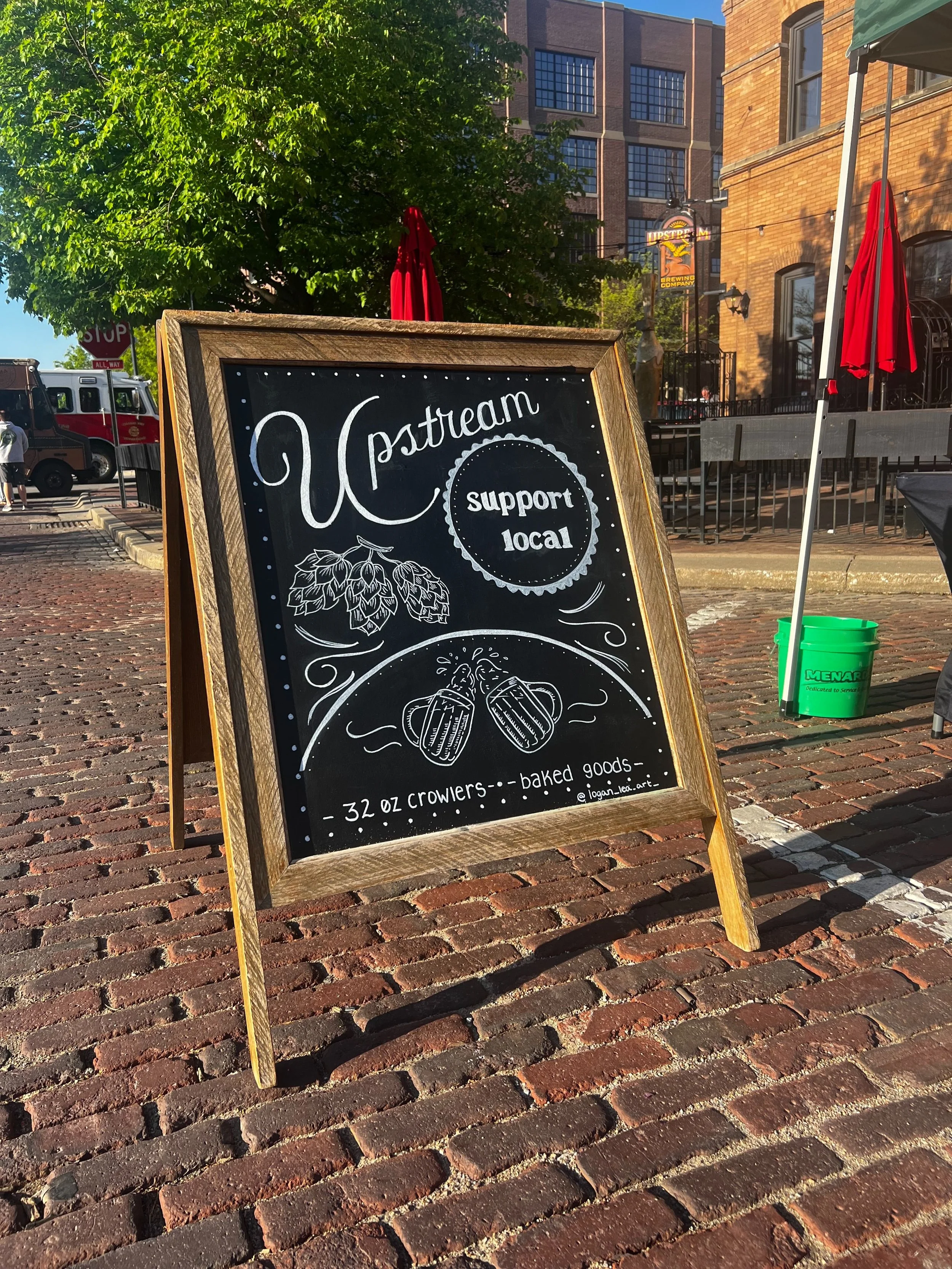 photo of a chalkboard sitting on cobblestones. Says upstream and support local. Drawing of hop plant and two beer glasses clinking. Bottom says 32oz crowlers and baked goods.