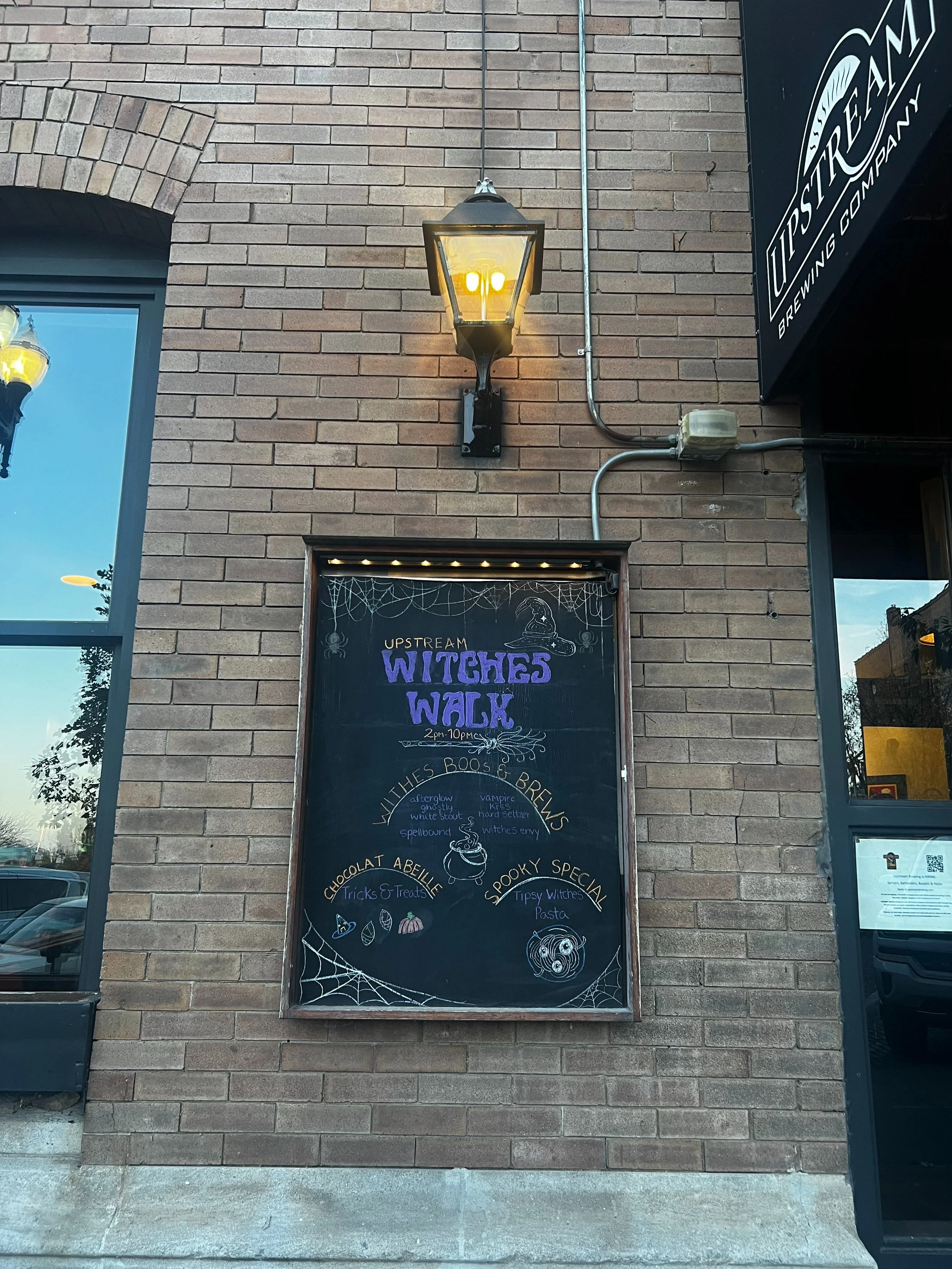 Chalkboard on a brick wall infront of a resturant. Says witches walk in purple with fun witchy drawings. Words that say Witches Boo Brews, Chocolat Abeille, and a Spooky Special. Spider web drawings at the top and bottom.