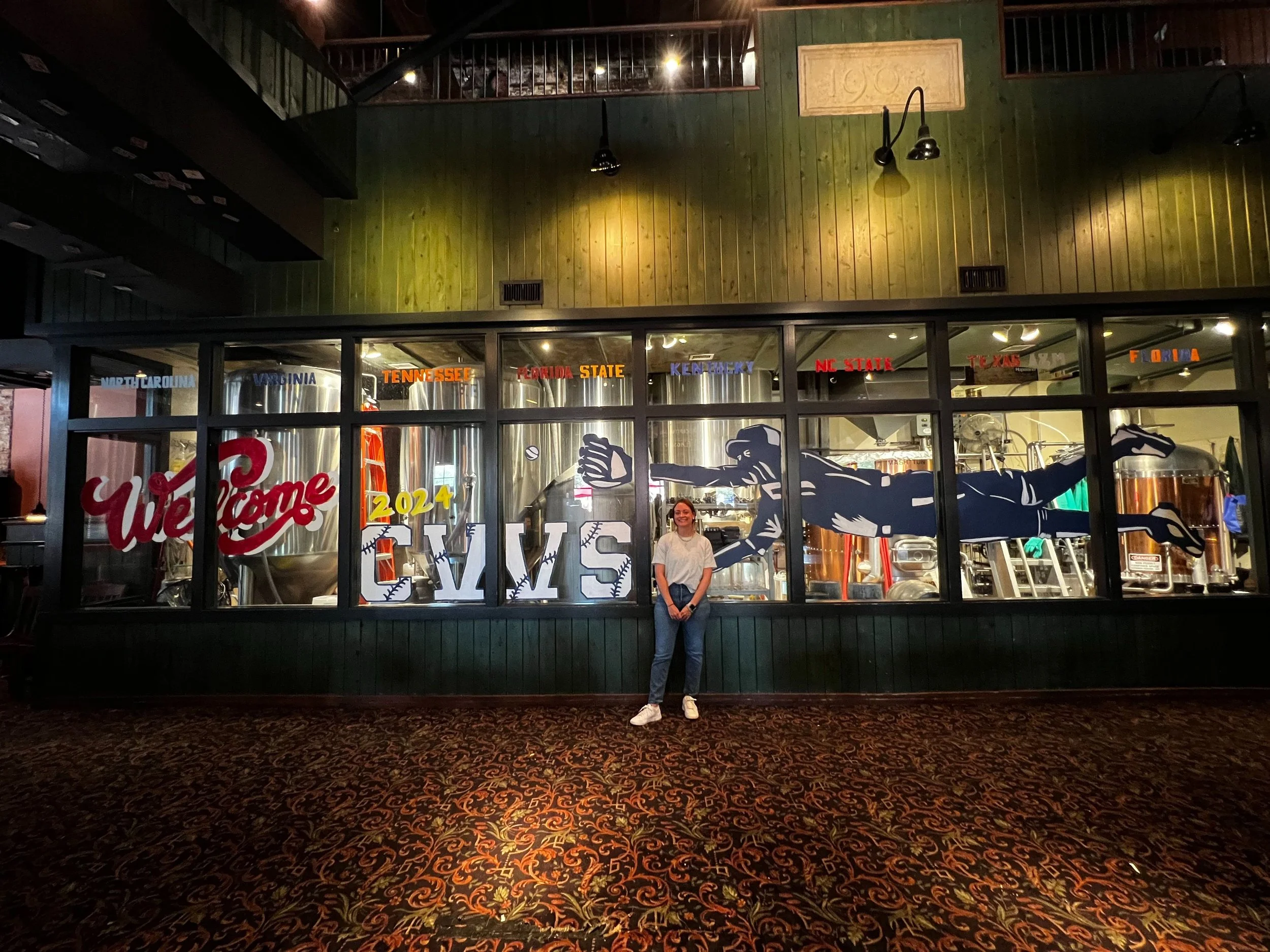 window art across 8 windows that says welcome 2024 cws with a silhouette painting of a baseball player catching a baseball. woman standing in front of it 