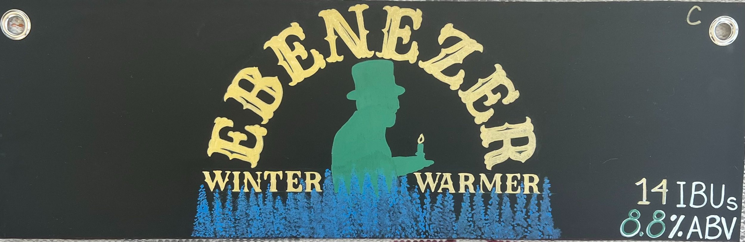 Chalk design that says Ebenezer in gold. With a green silhouette of an old man in a top hat holding a candle, it says winter warmer next to him in gold. Blue pine trees along the bottom. 