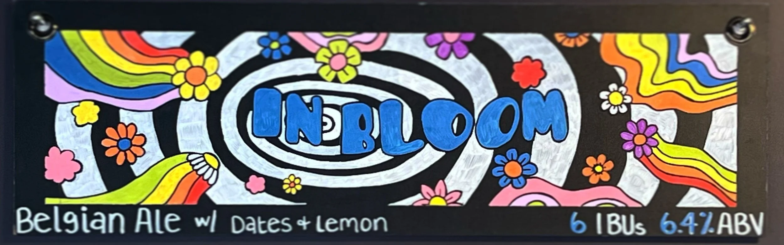Chalk art of the words "IN BLOOM" with funky floral rainbow designs with white stripes behind it.