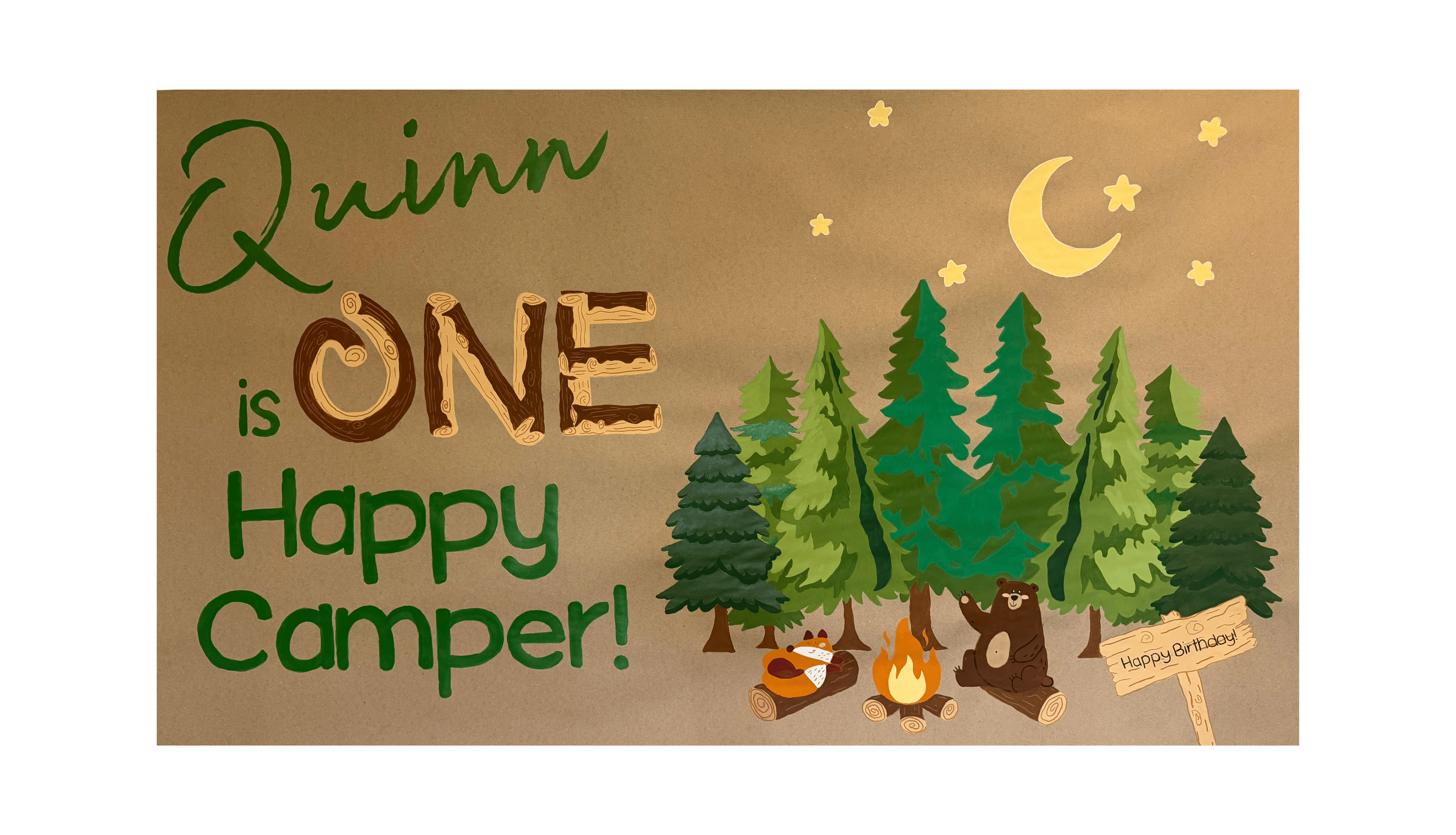 Banner art on brown craft paper the left says "Quinn is ONE Happy Camper" all in green except ONE looks like pieces of wood. To the right is a group of forest trees in multiple colors of green. In front of the trees are a bear and fox sitting on wood