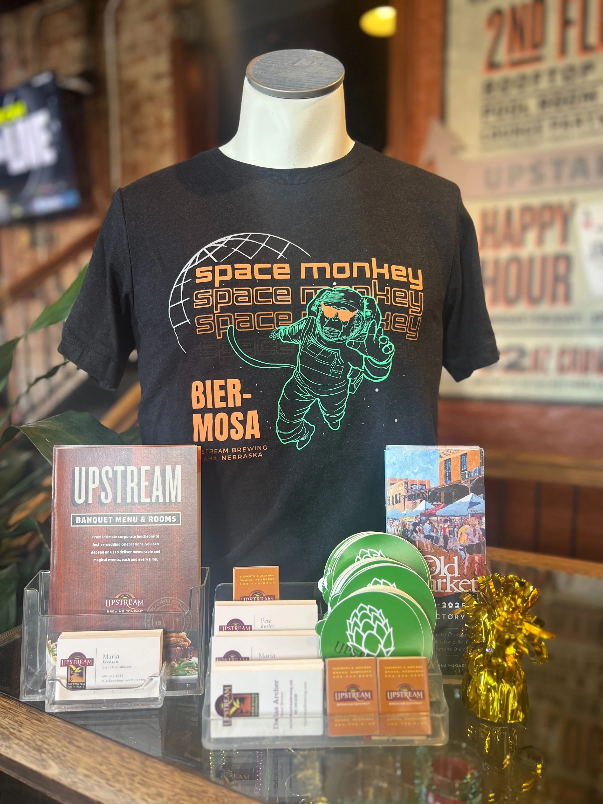 photo of a tshirt on a mannequin that says space monkey three times with a green monkey astronaut wearing orange sunglasses. says bier-mosa and upstream brewing omaha, nebraska.