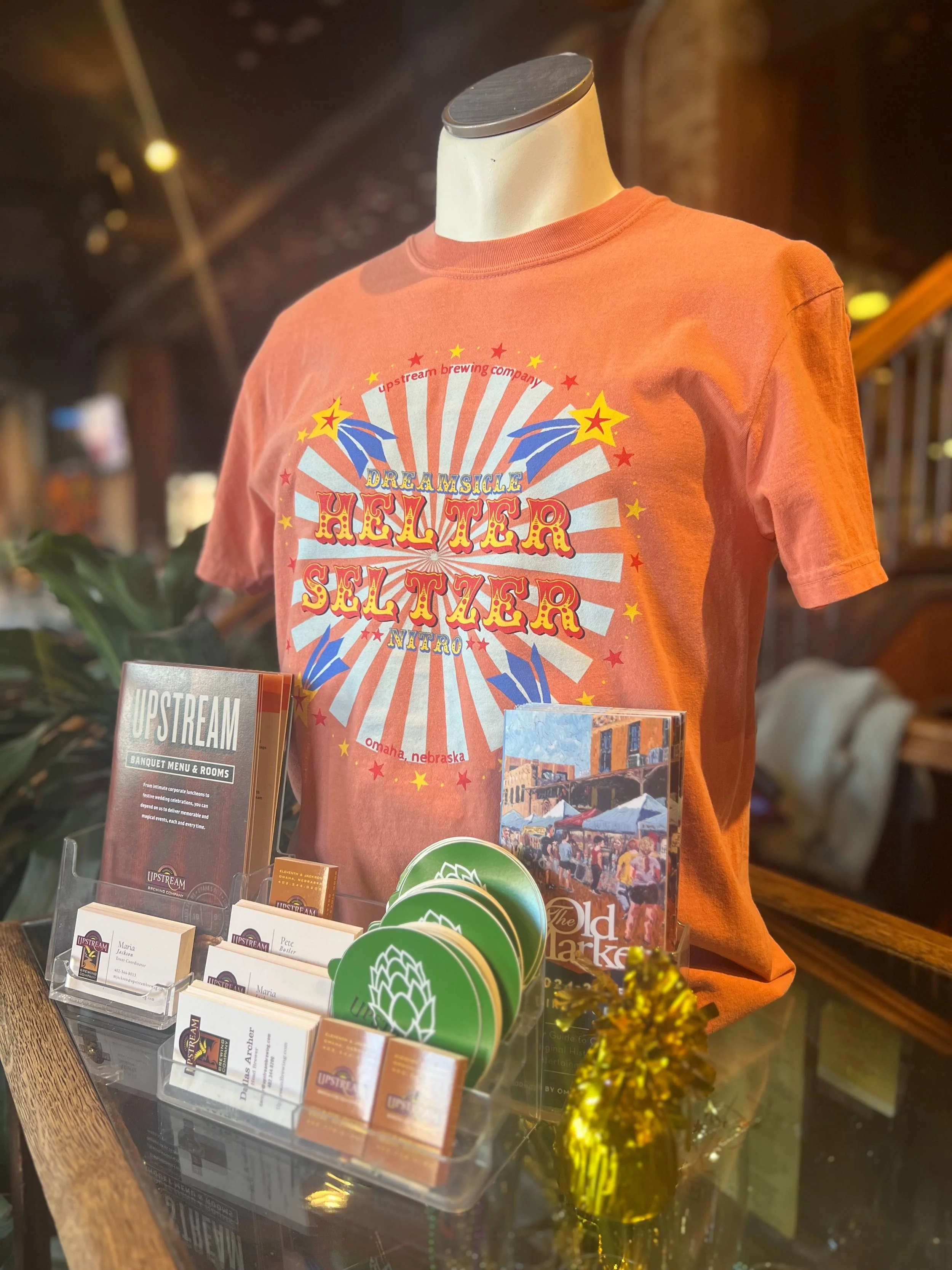 t-shirt on a mannequin orange color that says helter seltzer with a fun carinval design and shooting stars with blue and orange stripe circle behind it.
