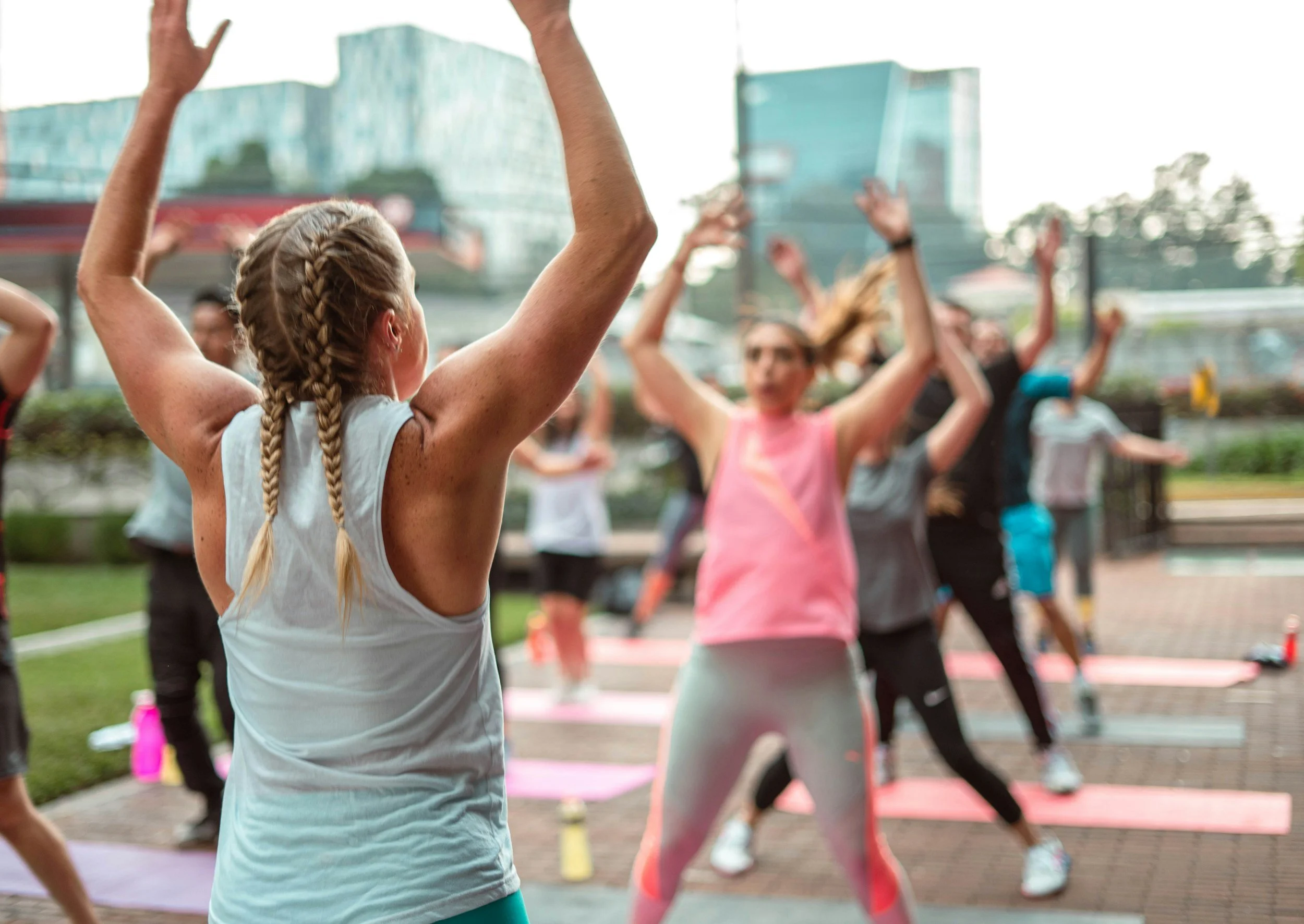 A group of women participating in an outdoor fitness class, following an instructor with arms raised, in a park with city buildings in the background.