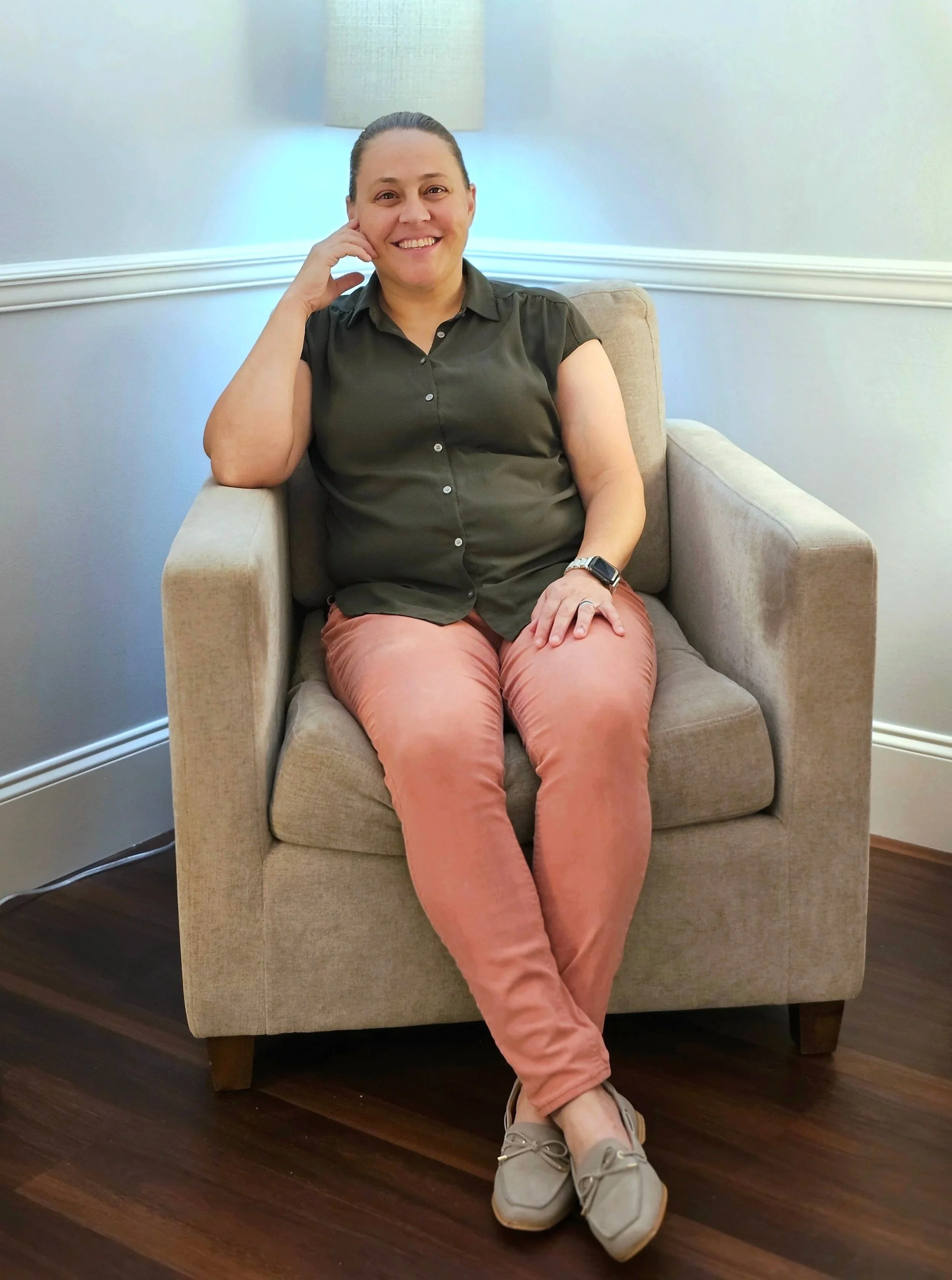 A smiling woman with her hair tied back, wearing a dark green sleeveless button-up shirt, rust-colored pants, light gray sneakers, and a smartwatch, sitting on a beige armchair in a room with gray walls, white crown molding, and dark hardwood floors.