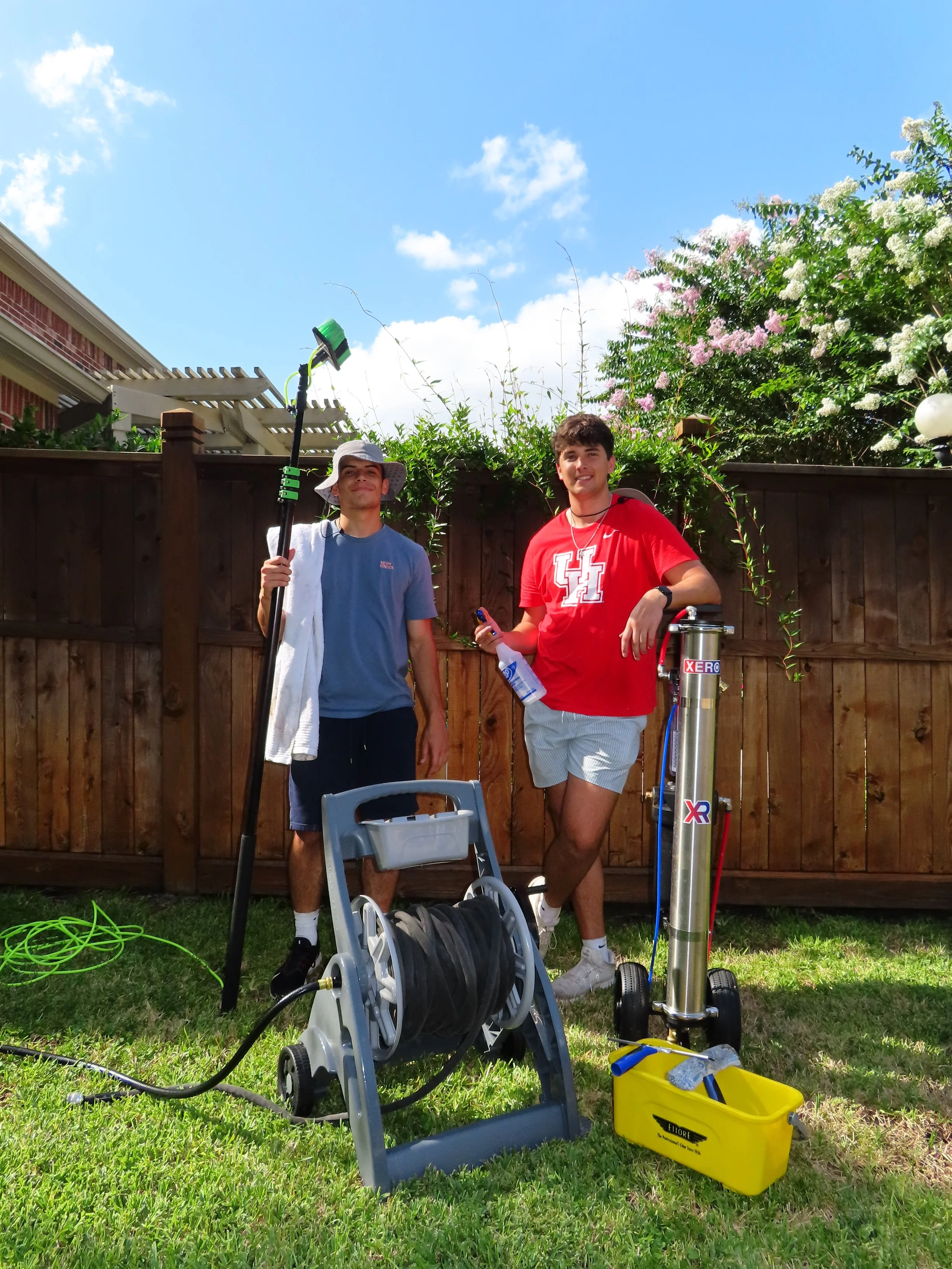 Two young men standing in a backyard, with one holding a long tool and the other holding a plastic bottle, surrounded by garden equipment including a hose reel, a yellow toolbox, and a water filtration system, with a wooden fence, flowering bushes, and a partly cloudy blue sky in the background.