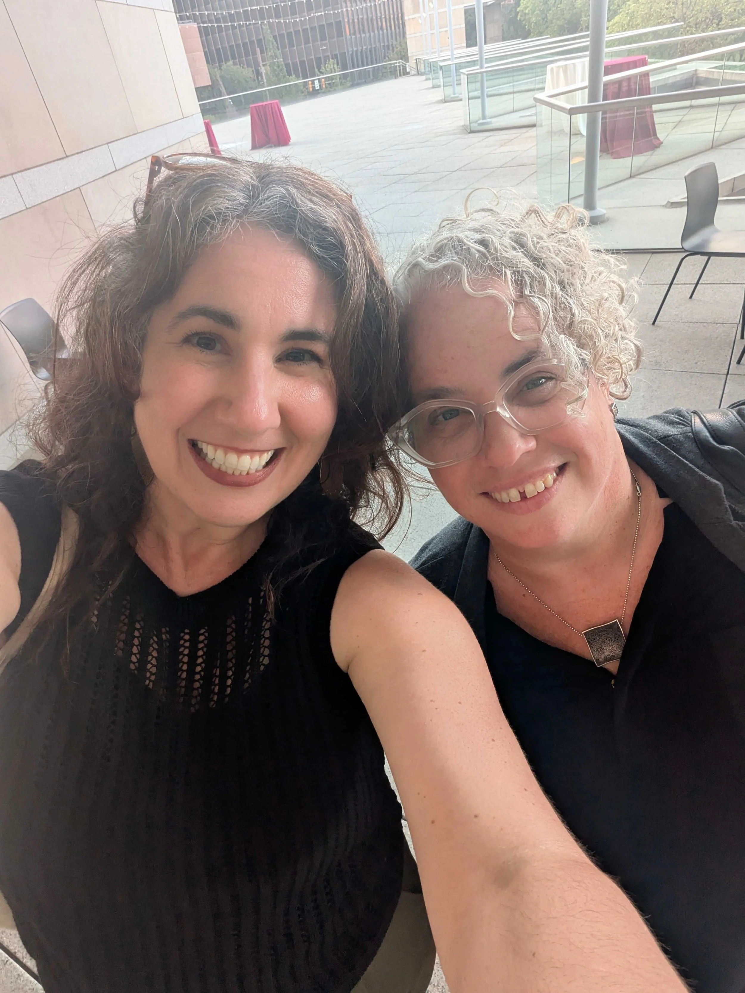 Two women smiling outside in a city landscape.