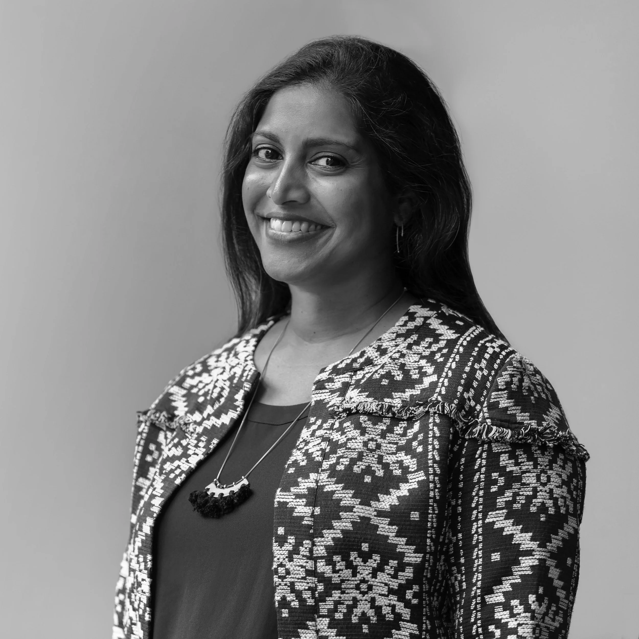 Black-and-white portrait of a smiling woman with shoulder-length hair, wearing a patterned blazer and a necklace, against a plain background.