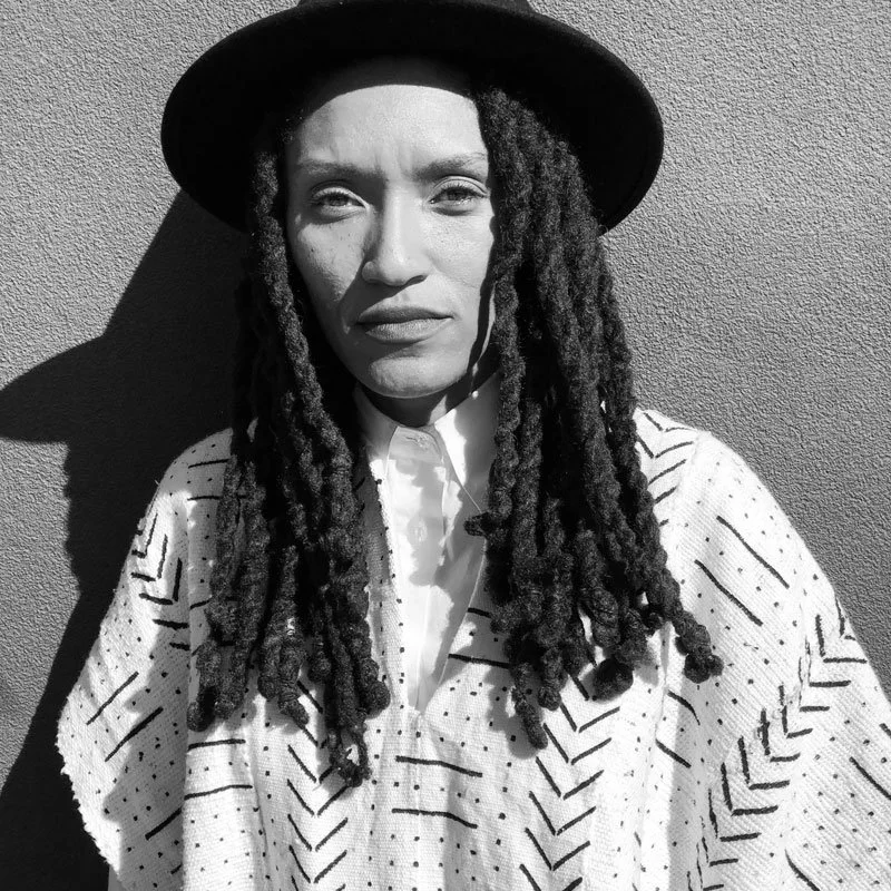 Black and white photograph of a woman with long dreadlocks wearing a wide-brimmed hat and a patterned shirt, standing against a textured wall.