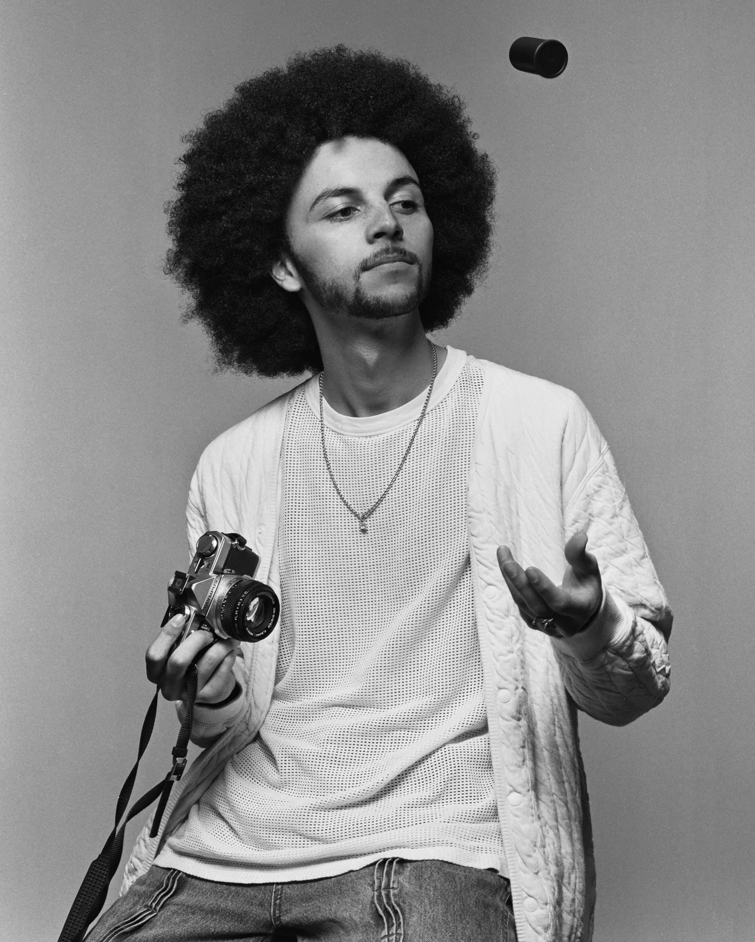 A black and white photo of a man with a large afro hairstyle, holding a vintage camera in his right hand and gesturing with his left. He's wearing a light-colored shirt, a patterned sweater, and a necklace, standing against a plain background.